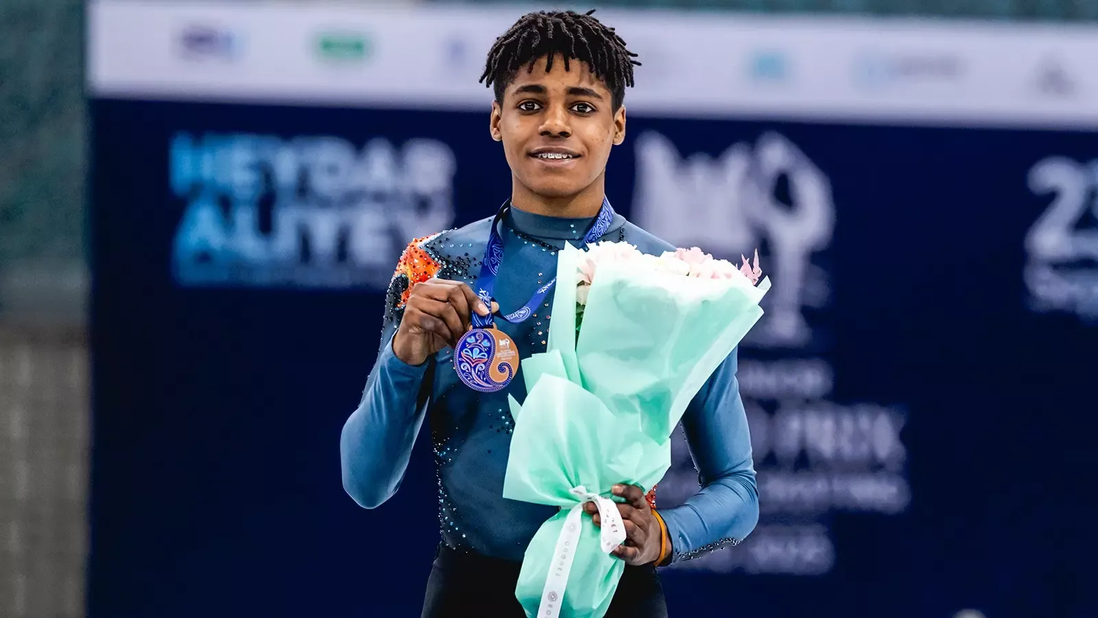 Caleb Farrington smiles while holding his bronze medal in one hand and a bouquet of flowers in the other. Caleb is a young Black man with braces and short dreadlocks wearing a dark teal top with orange detailing on the shoulder and black pants. 