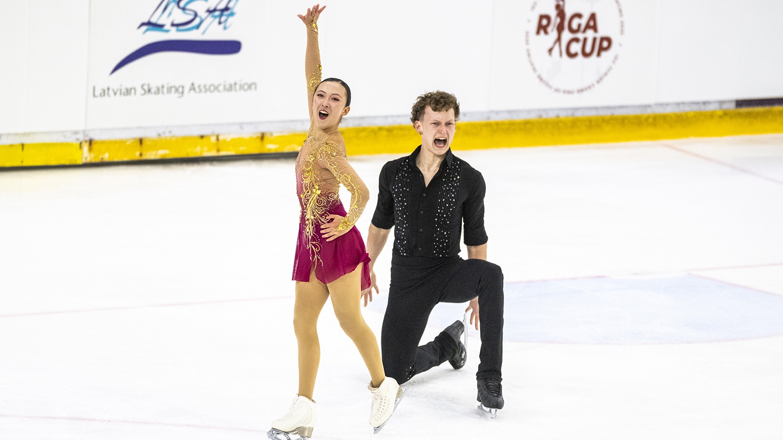 Pairs Naomi Williams and Lachlan Lewer deliver a powerful ending pose at JGP Latvia. Williams, dressed in red, has hand on hip and one extended into the air. Lewer, dressed in black, is kneeling on one knee with a powerful expression.