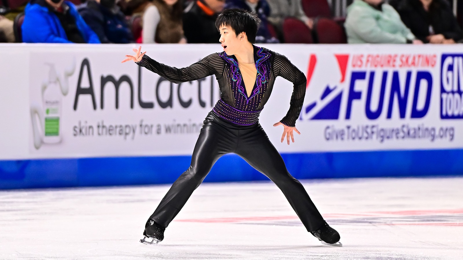 Tomoki Hiwatashi skates looking to the right with his right arm extended. He is a young man with short black hair wearing a black long sleeve top with a deep v neck and purple and blue beading around the collar. 