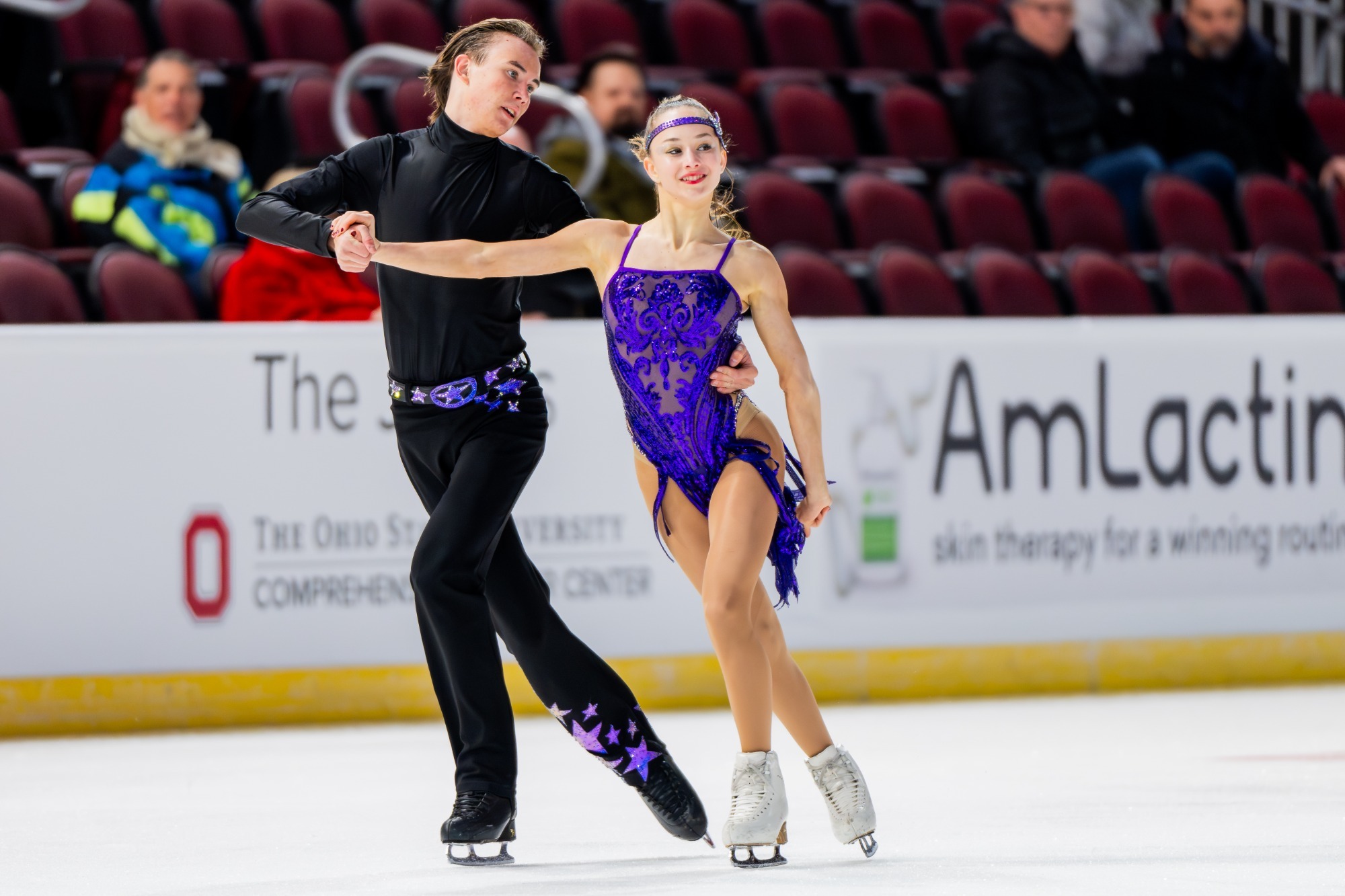 Dressed in all black, with purple accents on the waist and lower leg, Daniil Veselukhin skates hand in hand with Hana Maria Aboian, dressed in all purple.