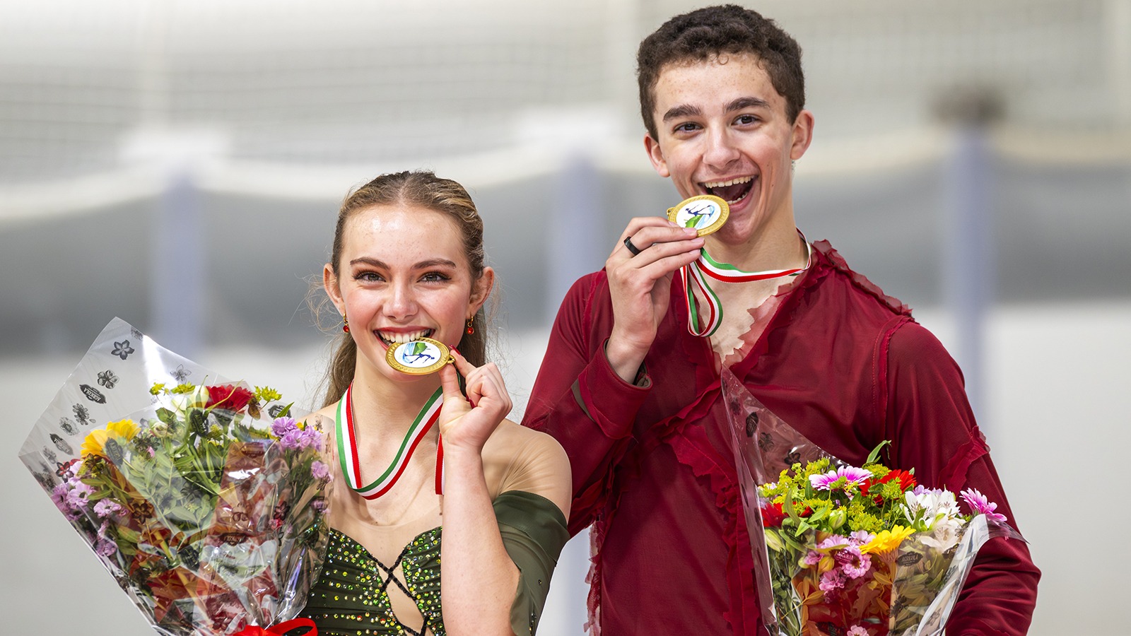 Jasmine Robertson and Chase Rohner stand on the podium biting their medals. They hold their medals in one hand and flowers in the other. Jasmine (left) is a young woman with long blonde hair weating a green skating costume. Chase (right) is a young man with short brown hair wearing a long sleeve red top