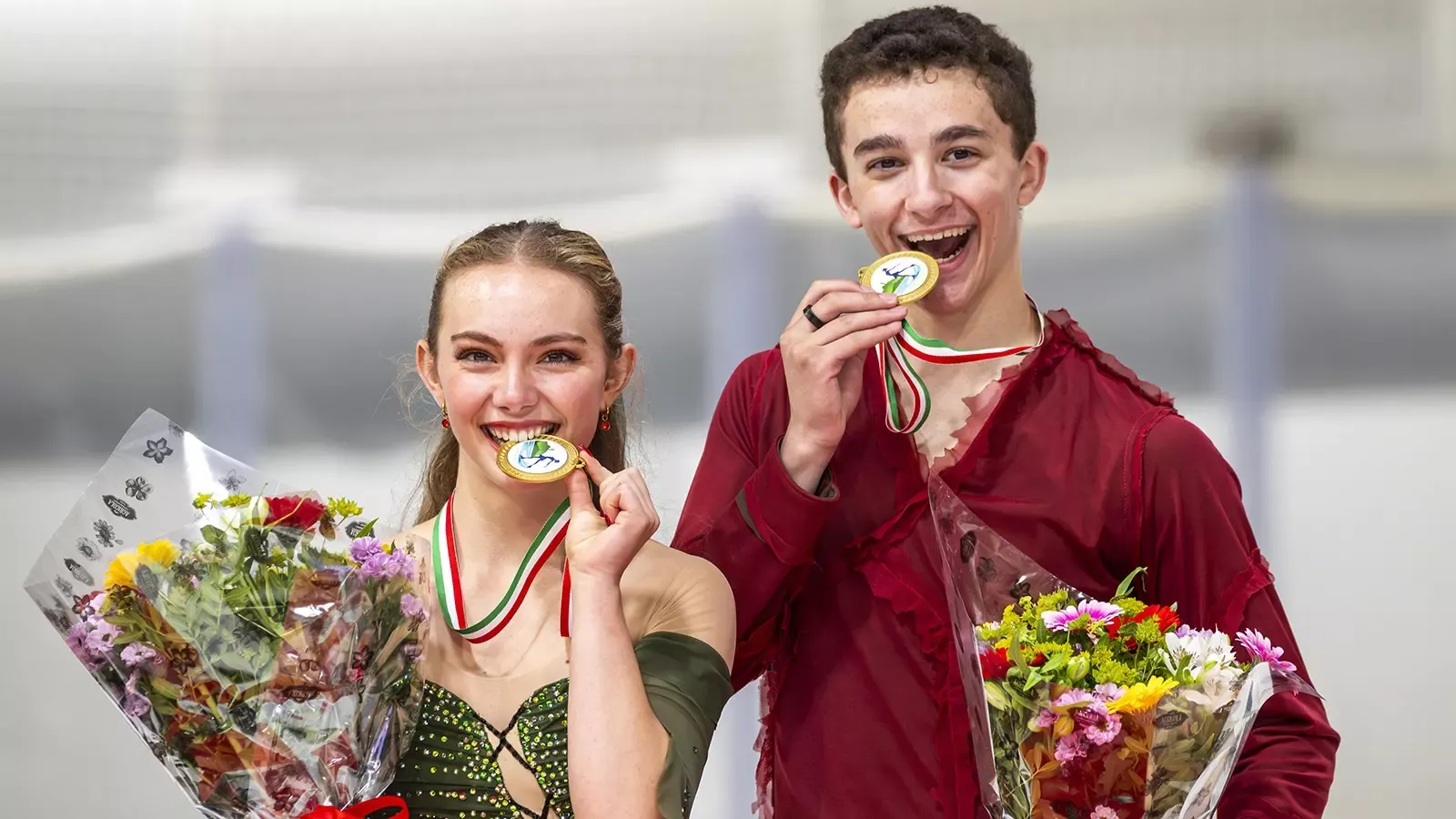 Jasmine Robertson and Chase Rohner stand on the podium biting their medals. They hold their medals in one hand and flowers in the other. Jasmine (left) is a young woman with long blonde hair weating a green skating costume. Chase (right) is a young man with short brown hair wearing a long sleeve red top