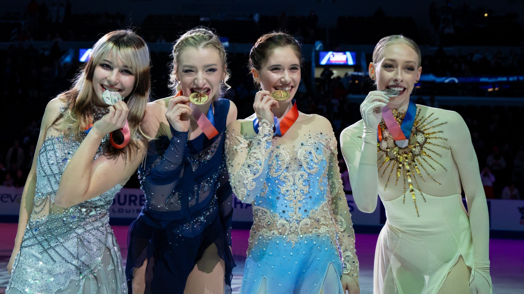 From left to right, Alysa Liu, Amber Glenn, Isabeau Levito and Bradie Tennell stand on the ice wearing their various free skate skating costumes. They all bite their medals as they look at the camera. 