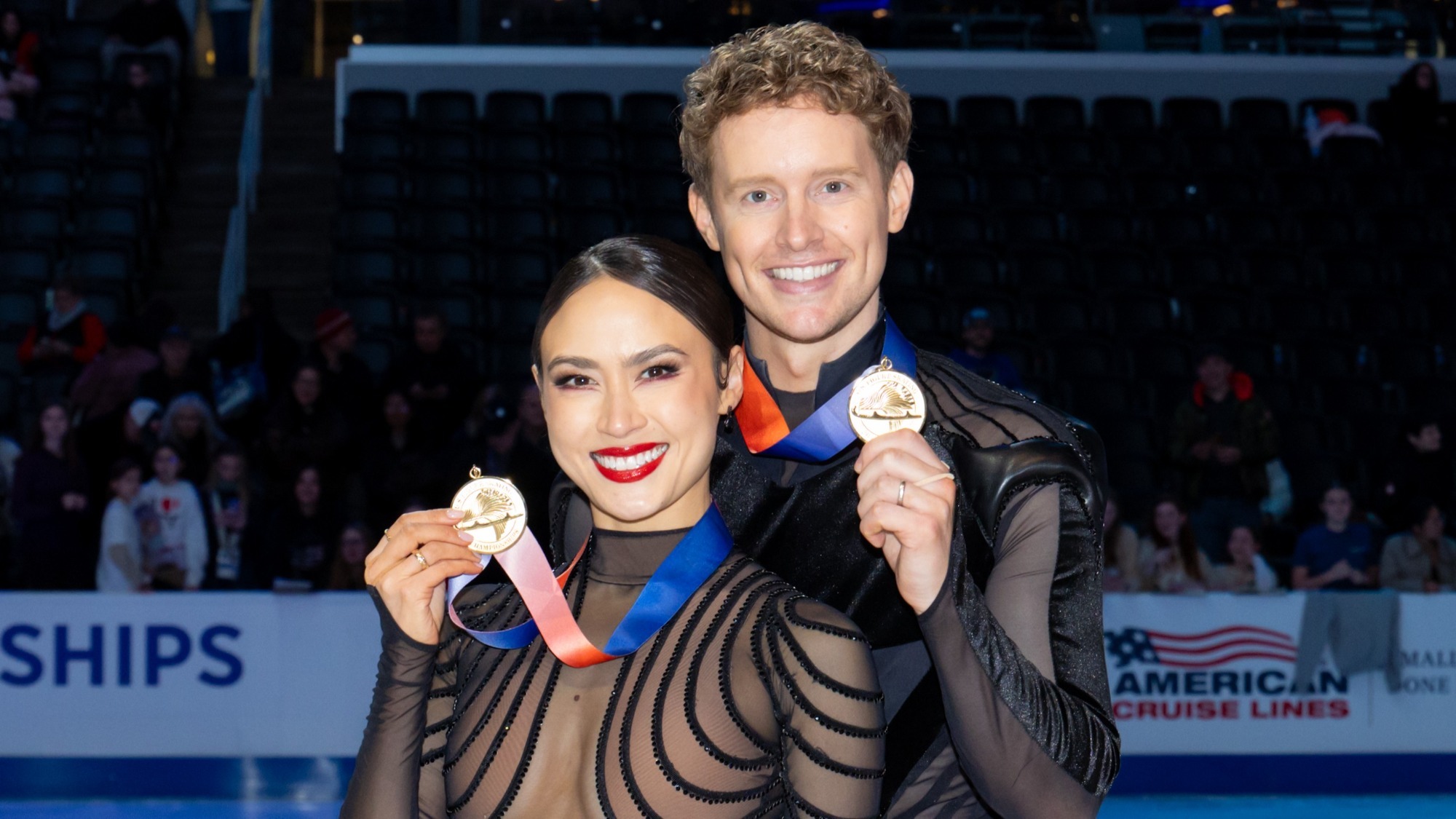 Madison and Evan Bates smile at the camera holding up their U.S. Championships gold medals which hang around their neck with a blue and red ribbon. Madison (left) is a woman with black hair tied back in a low bun wearing a black long sleeve dress with black stripes across the front. Evan (right) is a tall man with curly blonde hair wearing a black long sleeve top with sheer panels across the shoulders
