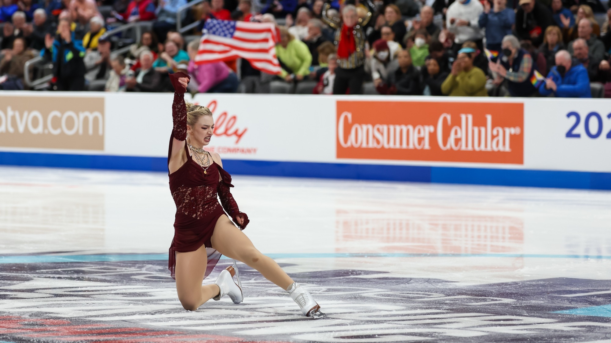 Amber Glenn kneels on the ice on one knee with her right fist raised in triumph. The crowd cheers holding American flags. Amber is a young woman with blonde hair tied back in a bun wearing a maroon skating costume with jewel detailing and off-the-shoulder sleeves