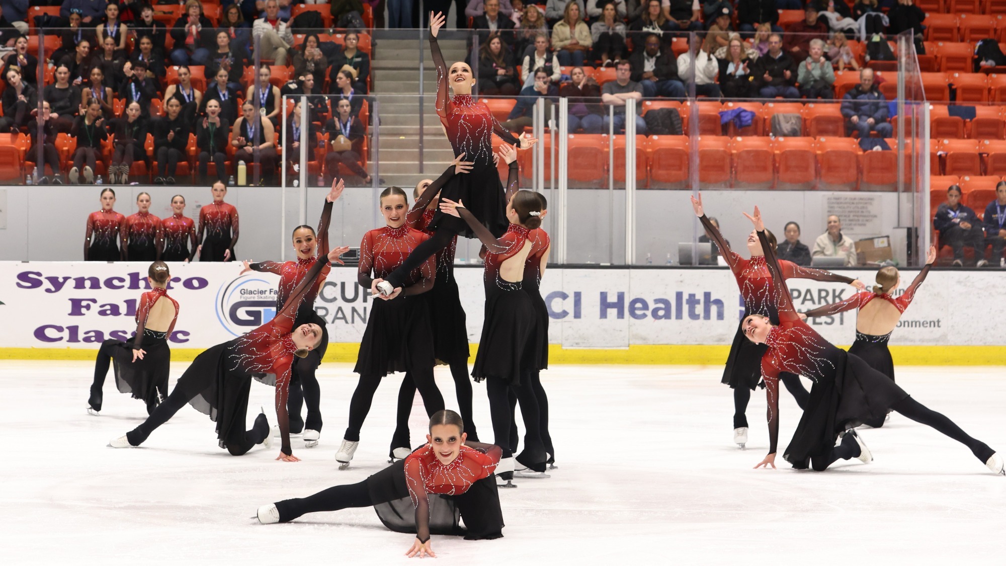 The Fond du Lac synchro team performs a lift with one skater being held up by four others at the base. Other skaters kneel or stand around them with one arm raised upward. They are all wearing matching long sleeve costumes with a red top, black skirt and silver jewel detailing down the torso and arms. A crowd of spectators looks on in the background. 
