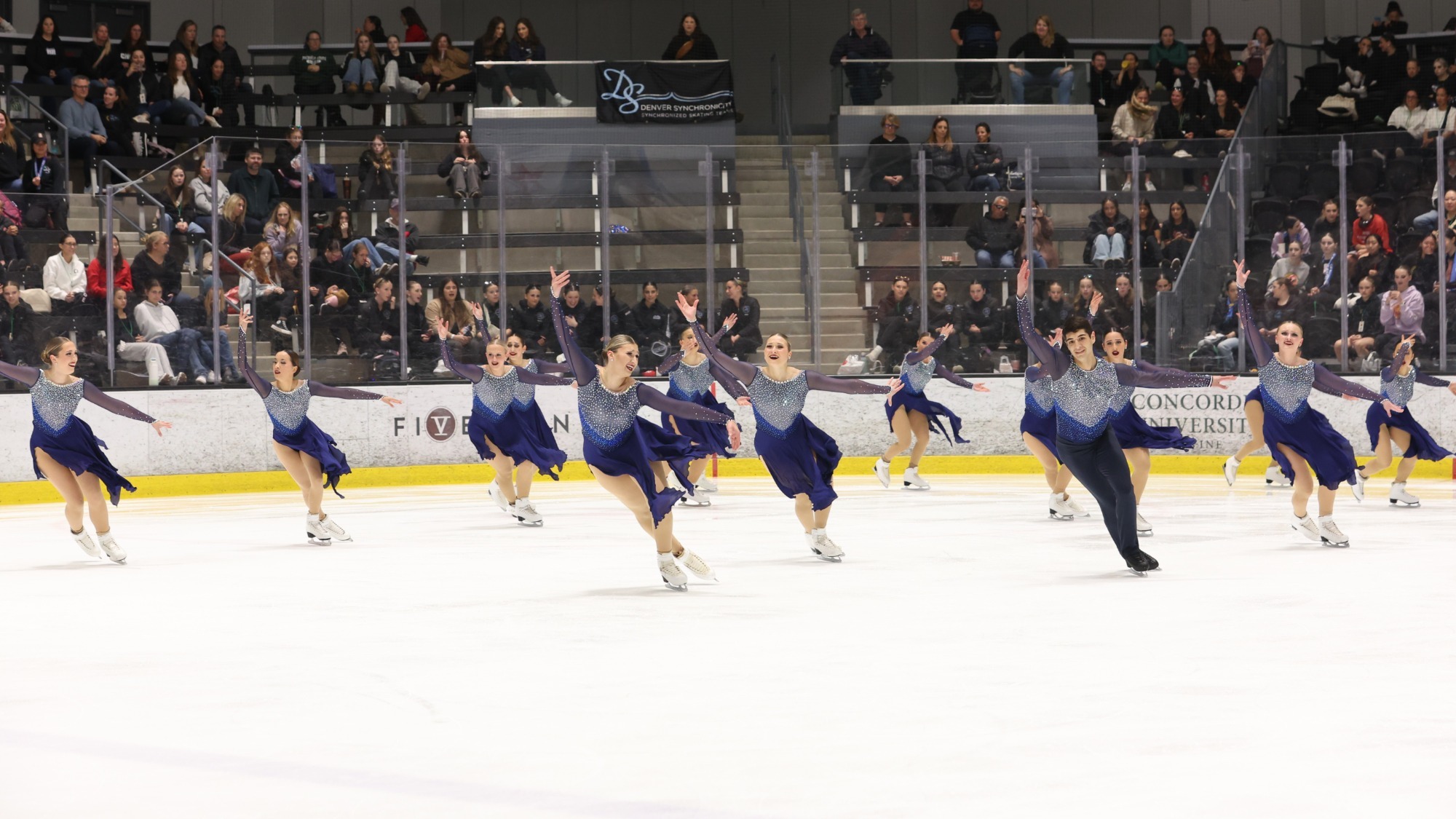 The Skyliners skate in a line with their right arms extended upward. They are all wearing matching dark blue skating costumes with a jeweled top, sheer sleeves and a flowy blue skirt. One male skater wears a matching costume with black pants