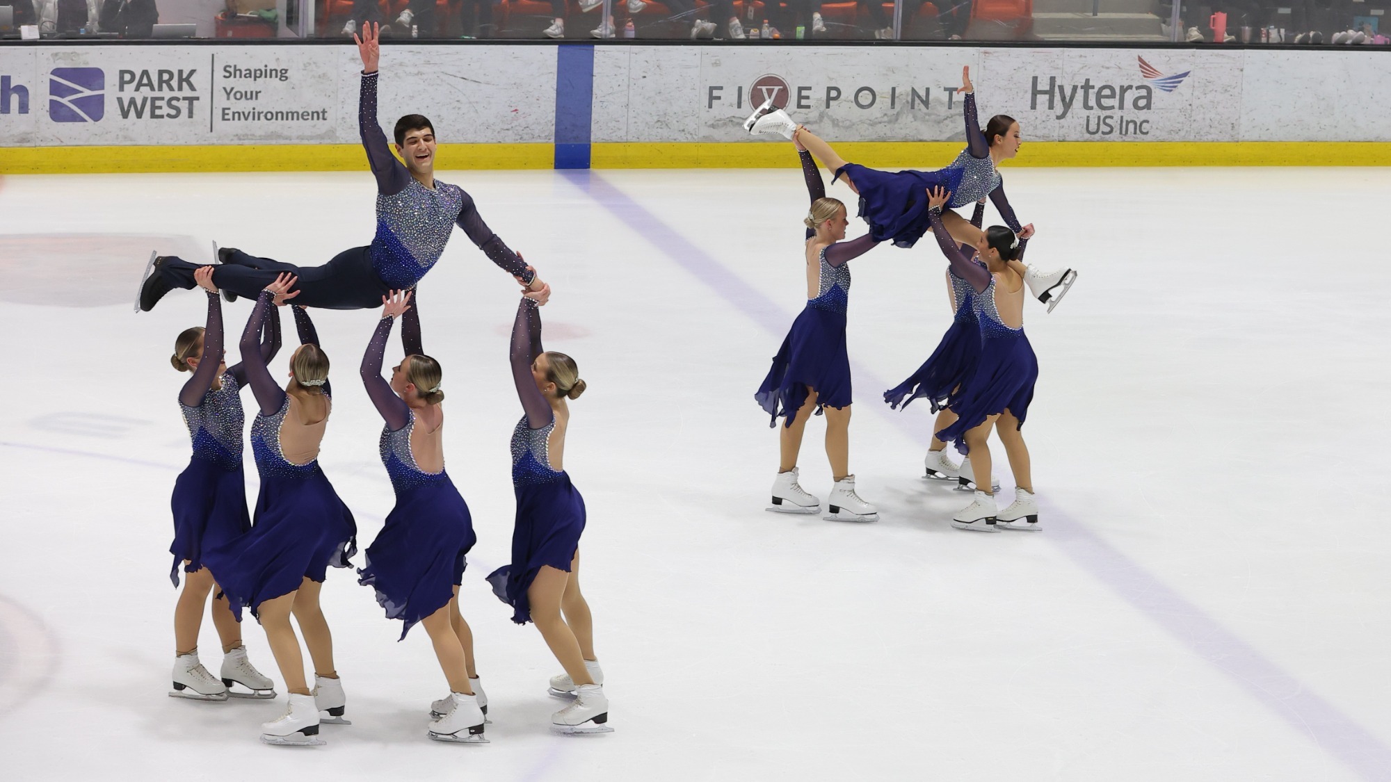 The Skyliners perform a group lift on the ice. On the ice, four women hold up one man, who is arched with his arm up. On the right, three women hold up another woman in a split. All wear navy blue with rhinestones.
