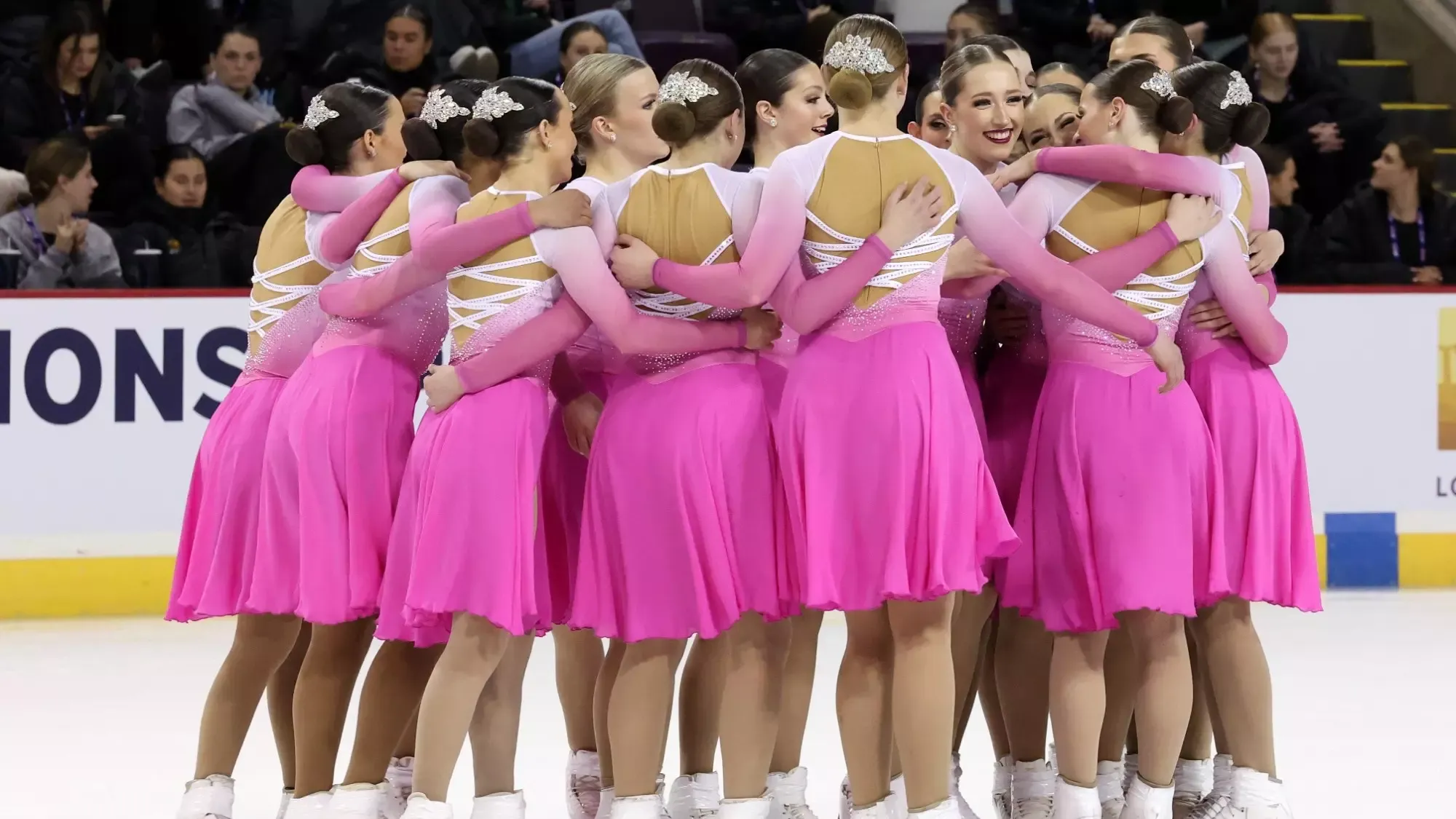 A synchro team wearing matching pink long sleeve skating costumes stands in a circle giving a group hug on the ice