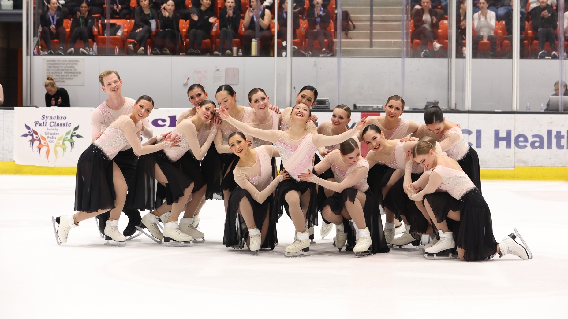 The Haydenettes stand together in a group with their arms around each other in a starting pose. They are all wearing matching skating costumes with a long sleeve light pink top and black flowy skirt. 