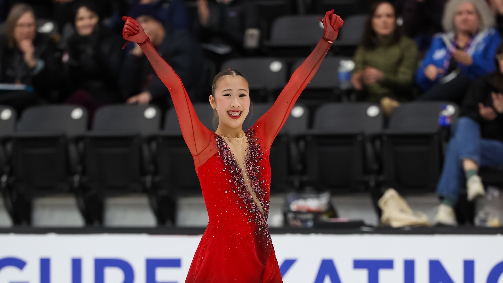 Hannah Kim smiles big with her arms raised above her head. She is a young woman with brown hair tied back into a bun and braces wearing a red long sleeve skating costume with matching red gloves