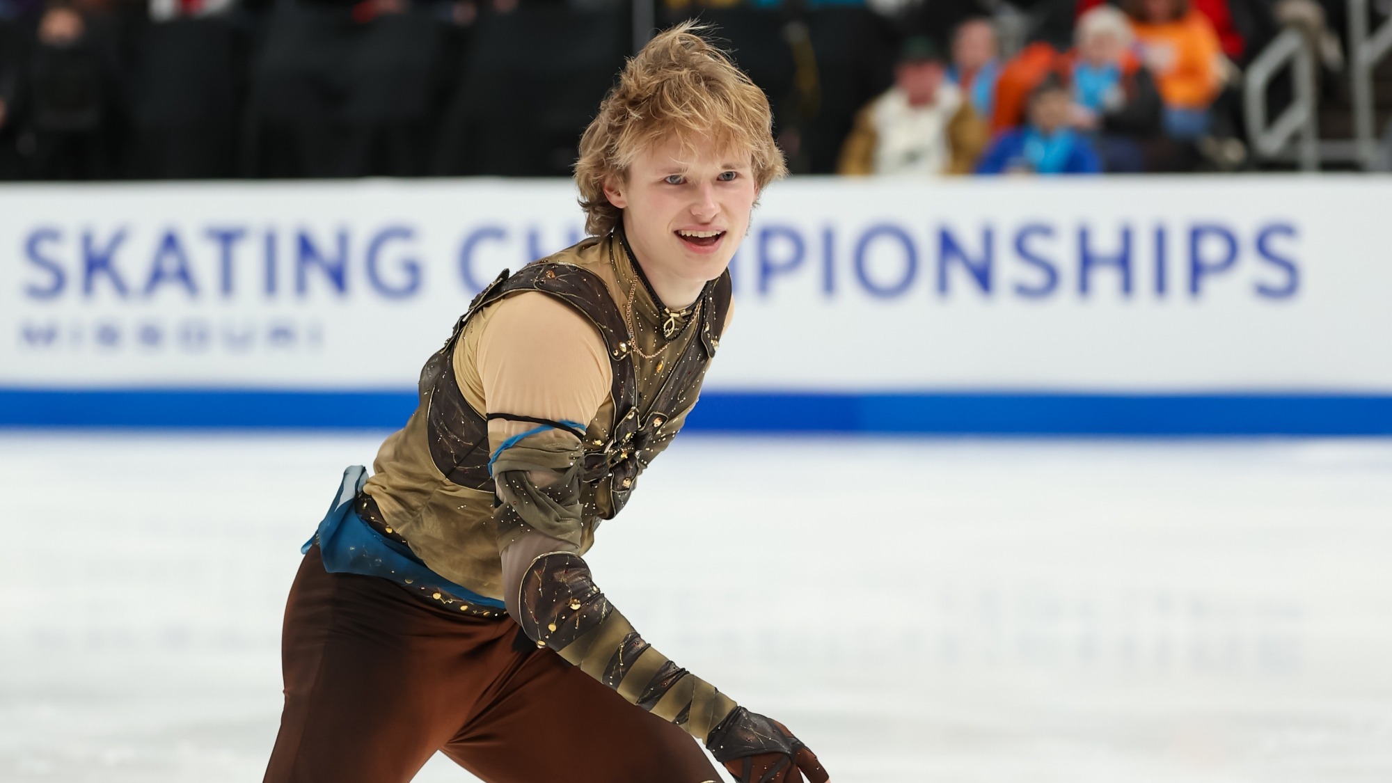 Ilia Malinin smiles at the camera during his performance. He is a young man with blonde hair wearing a tan long sleeve top with brown detailing, a blue belt and maroon pants
