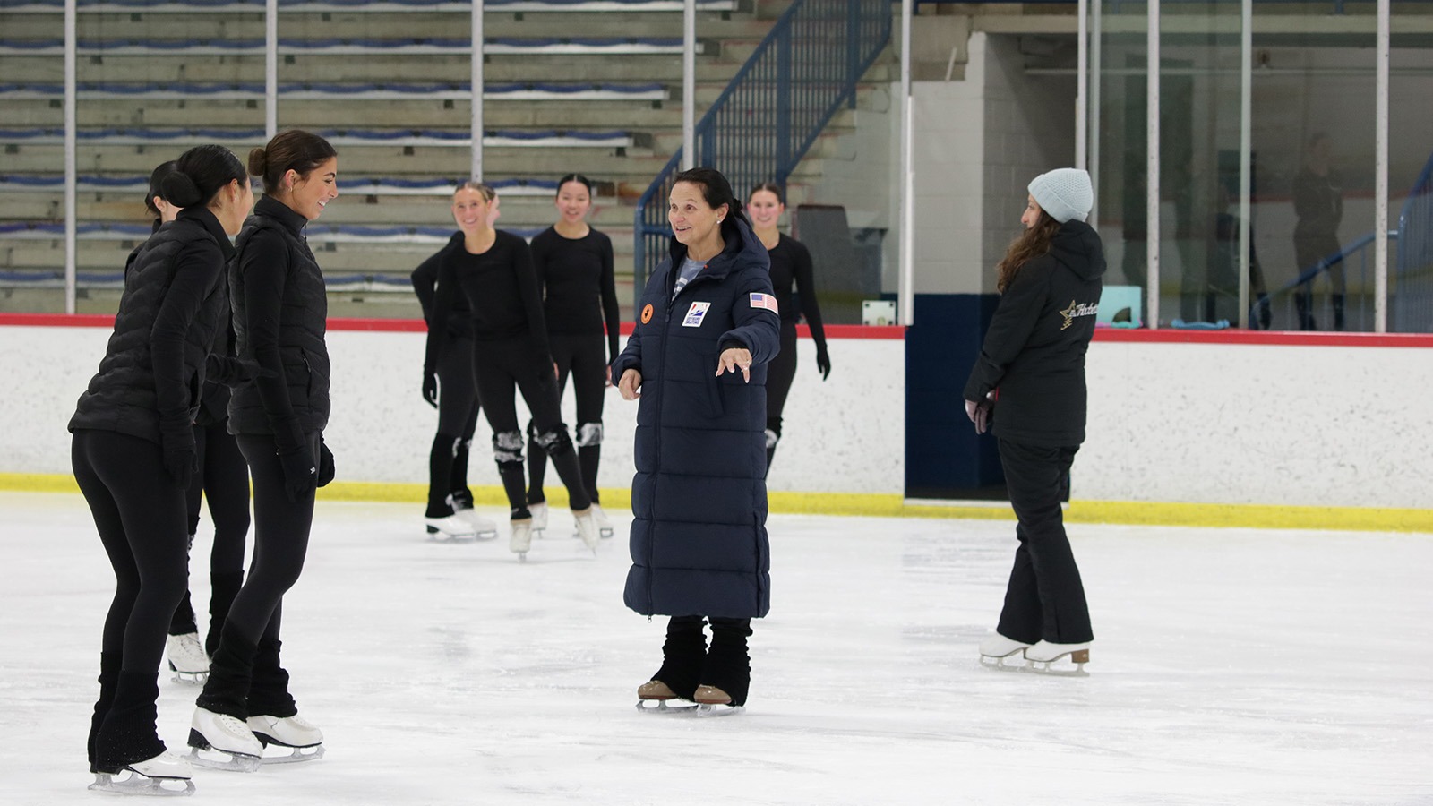 Pam May, dressed in a long blue U.S. National Team coat, directors members of the Hockettes on the ice.