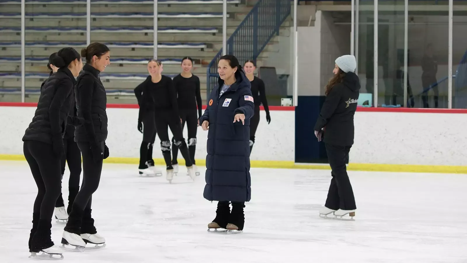 Pam May, dressed in a long blue U.S. National Team coat, directors members of the Hockettes on the ice.