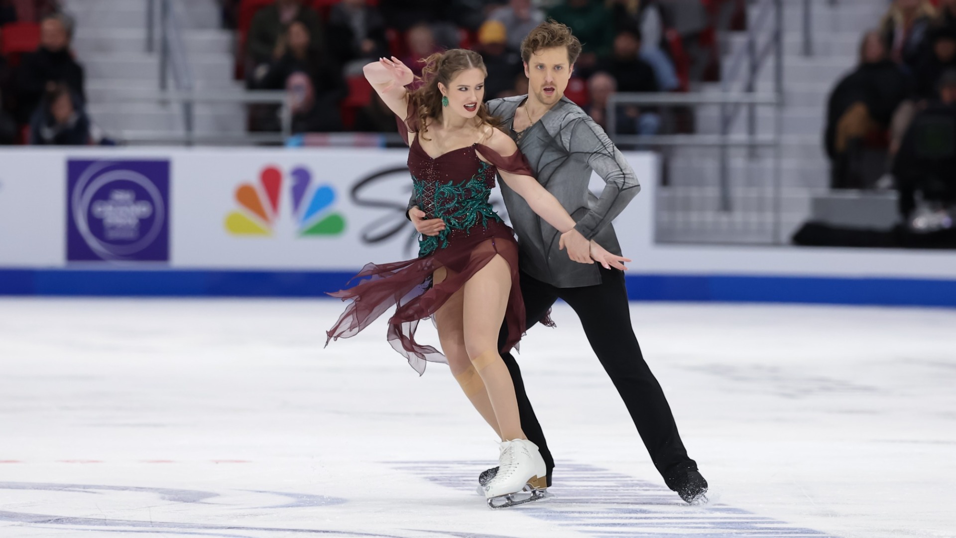 Christina Carreira and Anthony Ponomarenko perform at 2025 Skate America. Carreira wears a burgundy dress with sheer sleeves, flowing skirt panels and intricate teal embellishments, paired with white figure skates and teal earrings. Ponomarenko wears a gray, asymmetrical long-sleeved shirt with black geometric detailing and black pants. He holds Carreira around the waist as she extends one arm outward and raises the other near her head, both displaying expressive facial expressions.