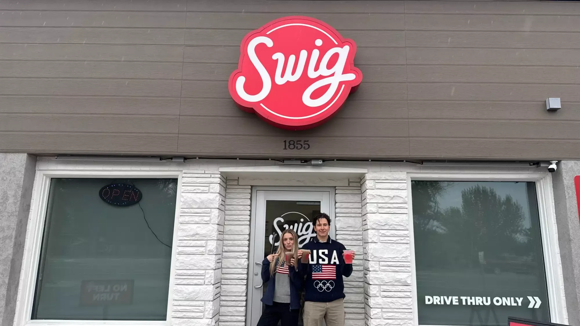 Emi and Vadym stand in front of a Swig store smiling and holding up their drinks. 