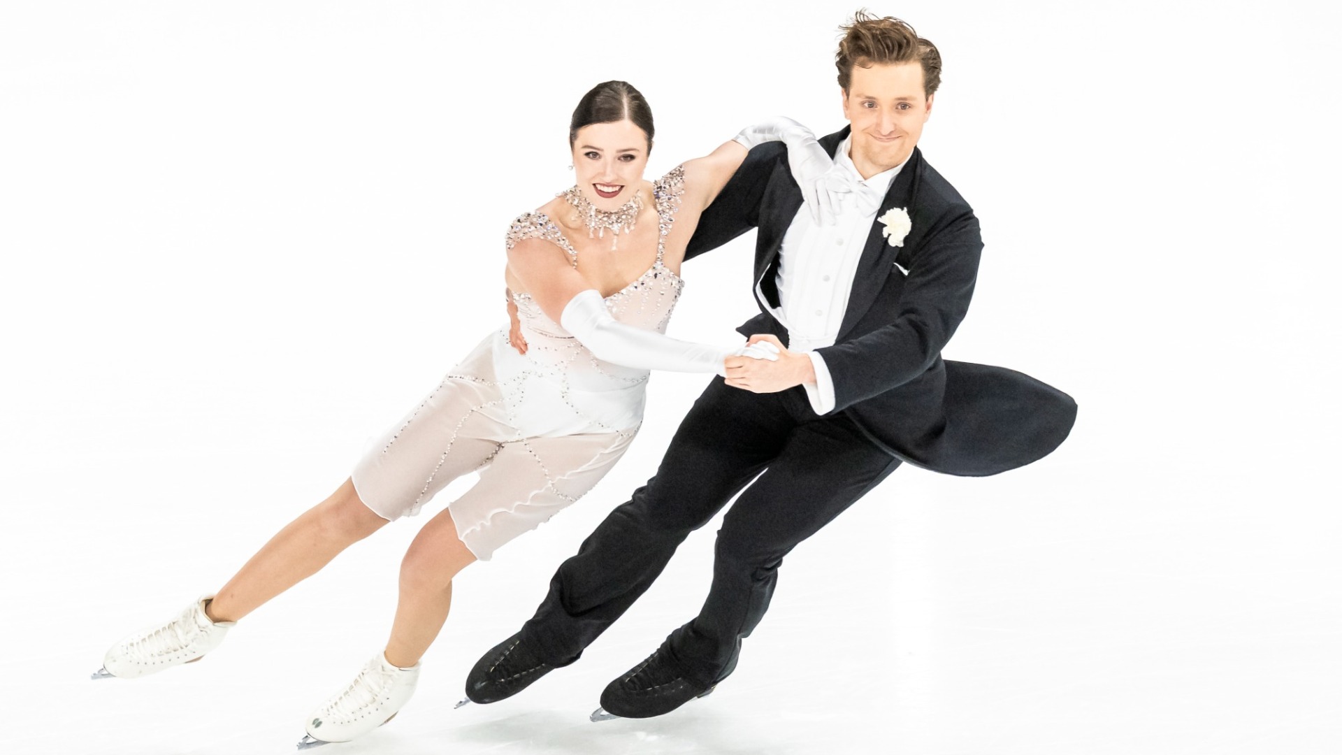 Christina Carreira and Anthony Ponomarenko perform an ice dance element on the ice. Carreira wears a white dress adorned with silver rhinestones and a matching choker, while Ponomarenko wears a black tailcoat with a white shirt, bow tie and white boutonniere. Both skate holding each other in a waltz position. 