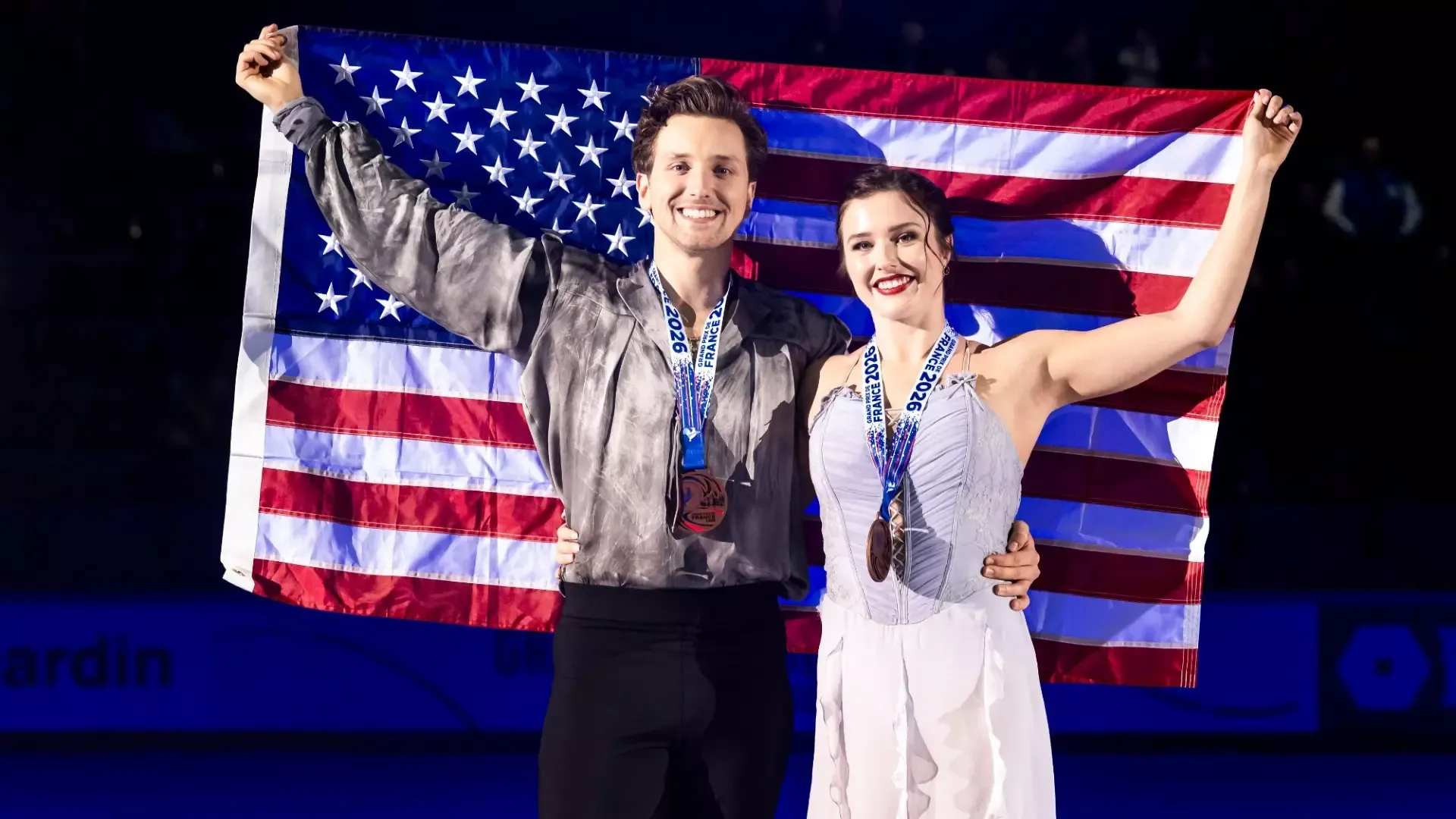 Christina Carreira and Anthony Ponomarenko stand side by side on the ice, smiling and holding an American flag stretched behind them. Both wear their Grand Prix de France 2026 medals around their necks. Ponomarenko wears a gray-and-black patterned top and black pants, while Carreira wears a grey skating dress. Both raise one arm to hold the flag
