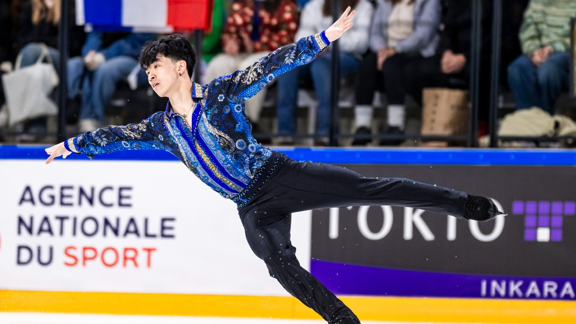 Jimmy Ma lands a jump with both arms extended and one leg stretched behind him. Jimmy is an Asian man with short black hair wearing a blue and black patterned long-sleeved shirt with intricate floral designs, silver and gold accents and black pants.