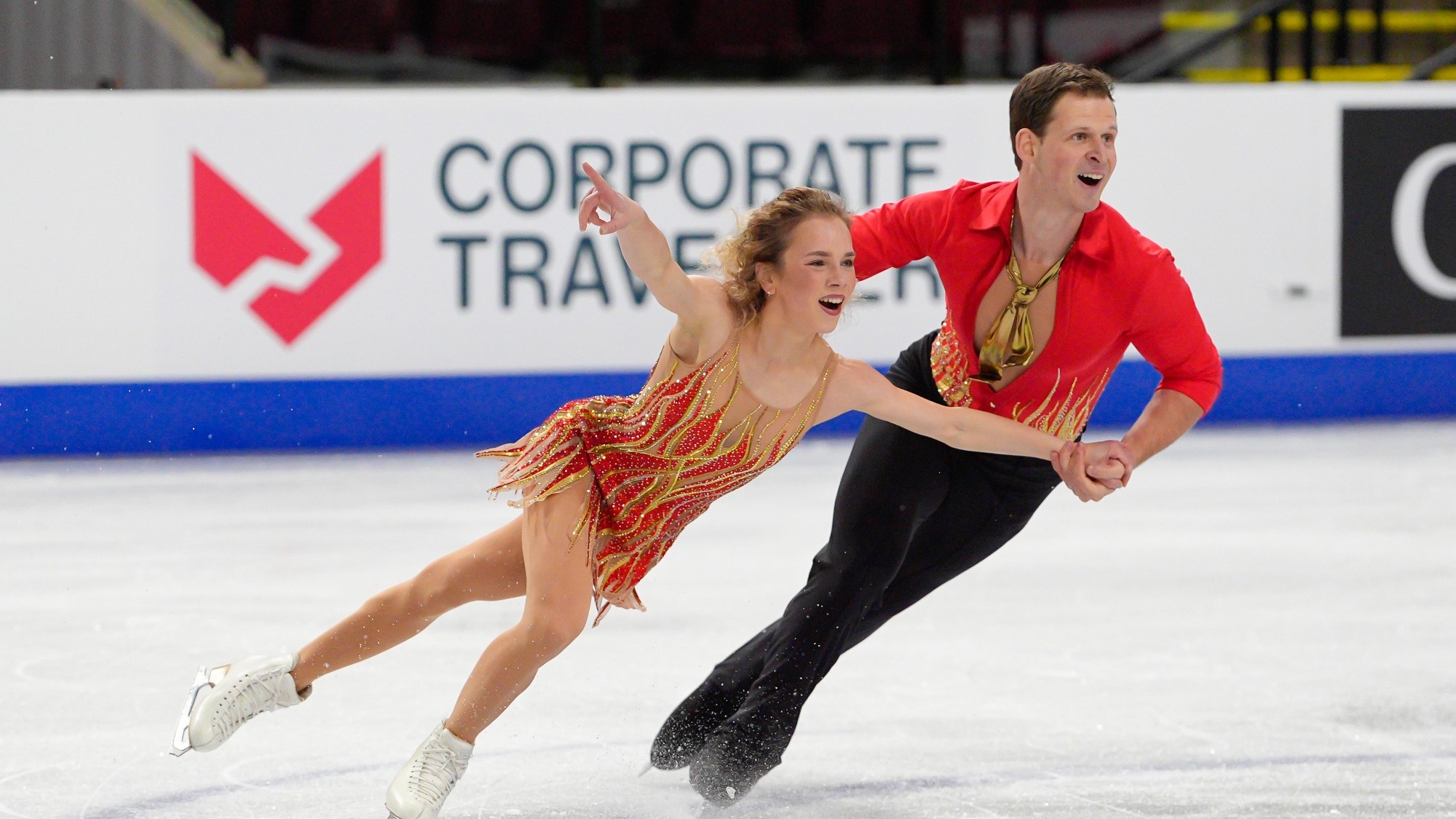 Alisa Efimova and Misha Mitrofanov skate their short program, The Last Dance. Efimova is wearing a dress patterned with yellow, orange, and red rhinestones shaped like flames. Mitrofanov is wearing a persimmon colored shirt with rhinestone accents, and black dress pants. 