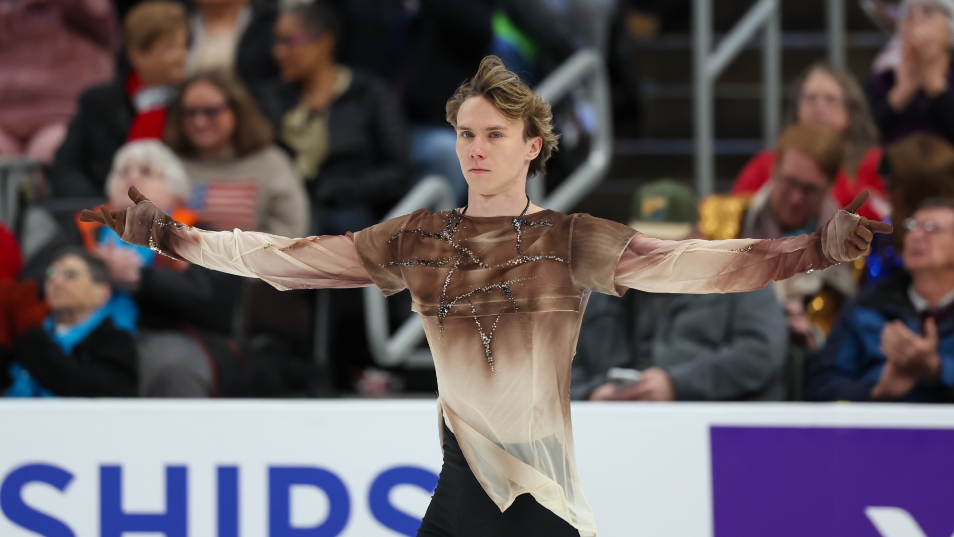 Daniel Martynov performs on the ice with his arms extended to either side, wearing a sheer beige costume embellished with rhinestones and black pants. Spectators watch from the stands in the background.
