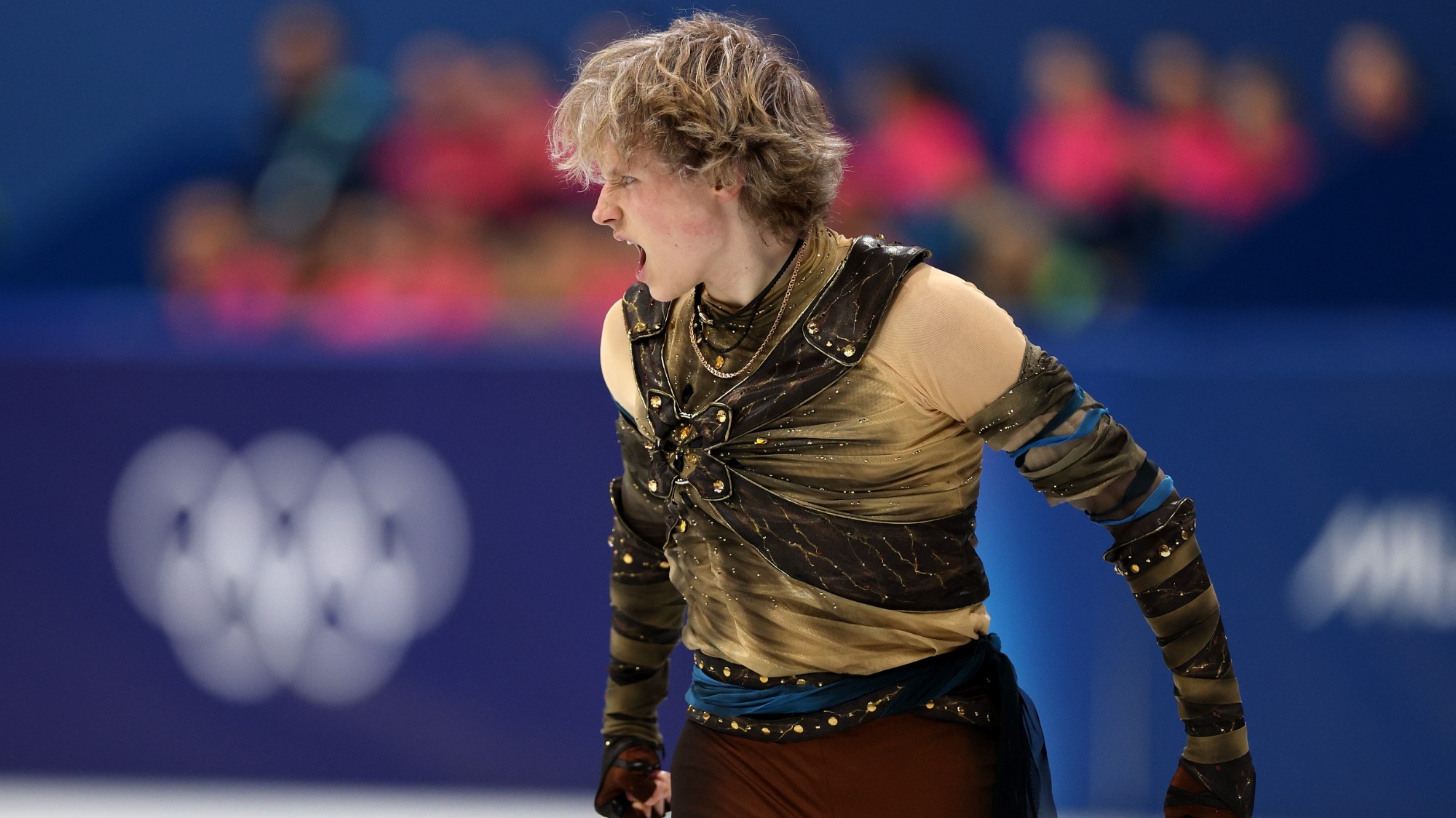 Ilia Malinin looks to the left with a look of determination on his face during his short program at the 2026 Olympic Winter Games. Ilia is a young man with short blonde hair wearing a brown long sleeve top with jewel details, a blue belt and matching brown pants. 
