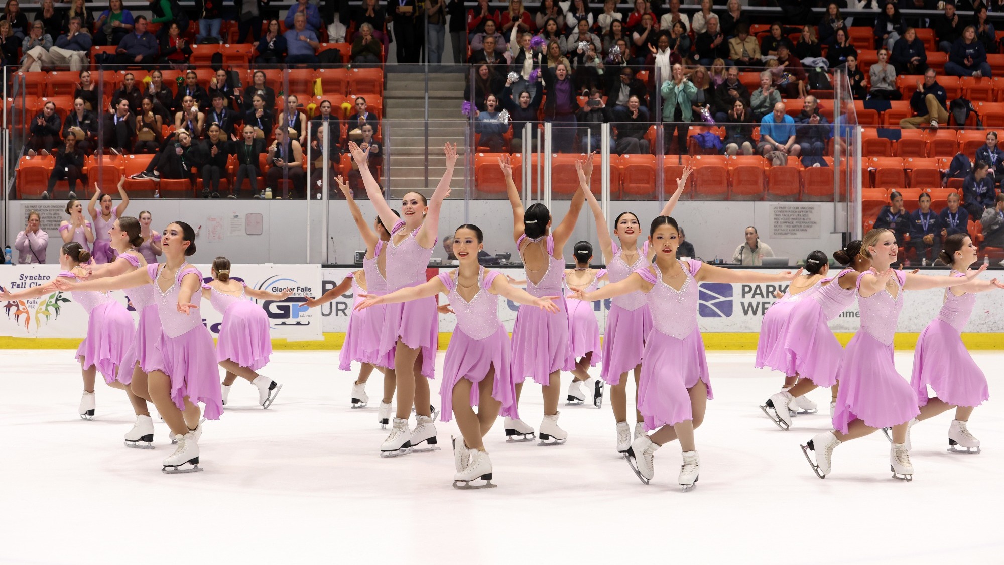 DC EDGE skates with their arms raised in unison wearing matching pink skating costumes