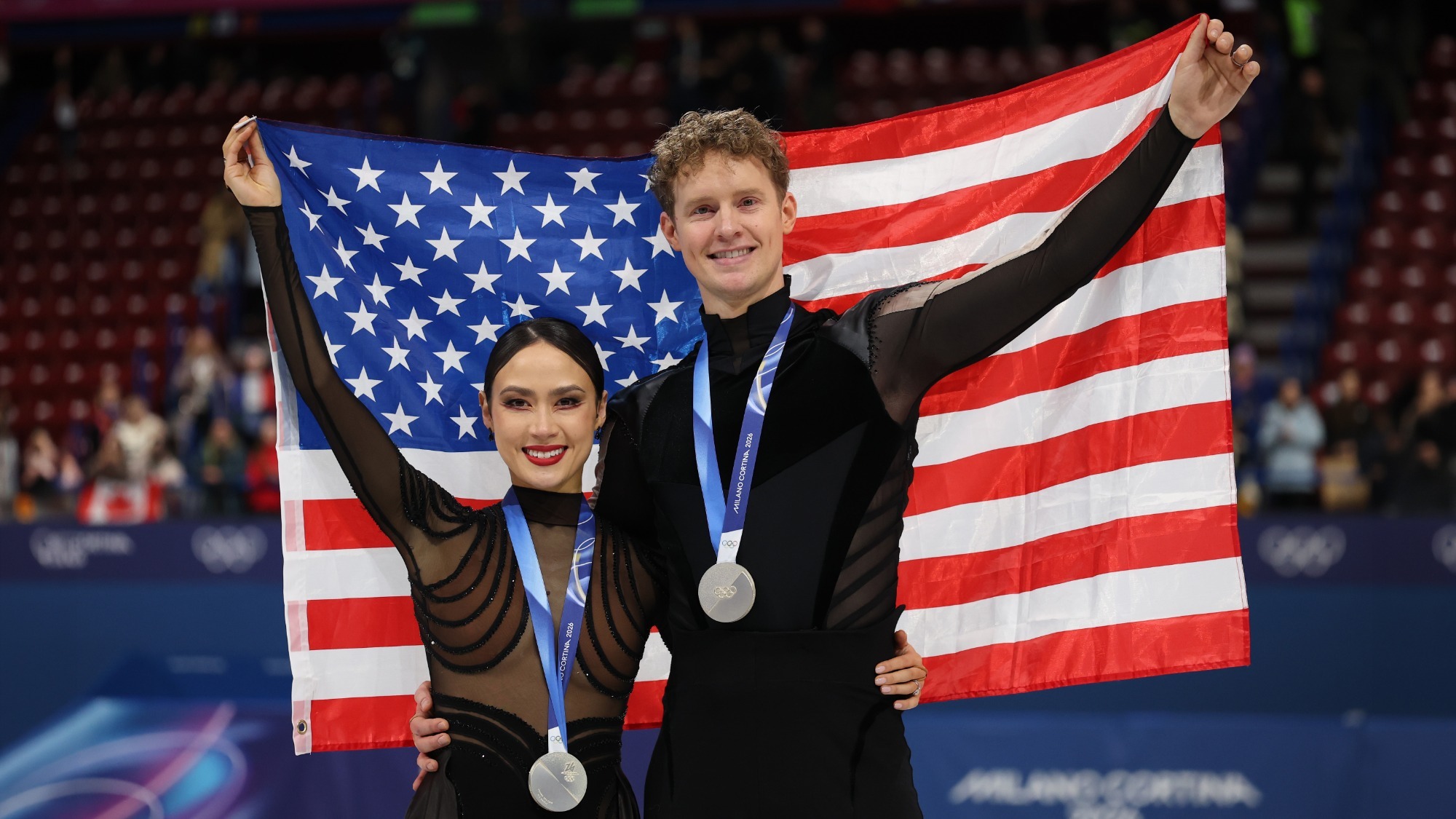Madison Chock and Evan Bates stand holding an American flag with their Olympic gold medals around their necks. Madison (left) is a woman with dark hair slicked back in a low bun wearing a long sleeve black costume. Evan is a man with curly blonde hair wearing a long sleeve top with a black torso and sheer brown sleeves. 