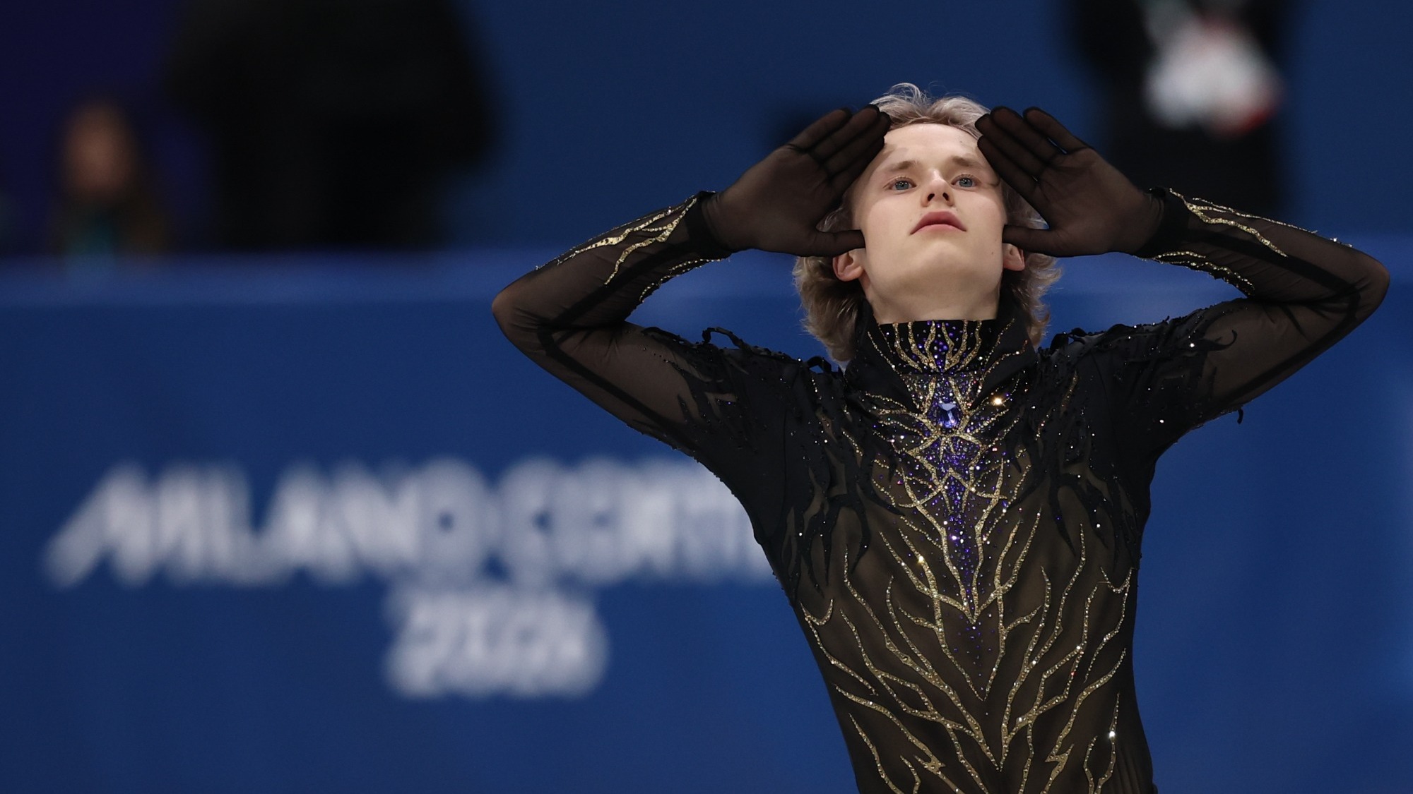 Ilia Malinin stands looking outward with both hands on his face. He is a young man with blonde hair wearing a long sleeve skating costume with gold and blue jewel detailing down the front. 