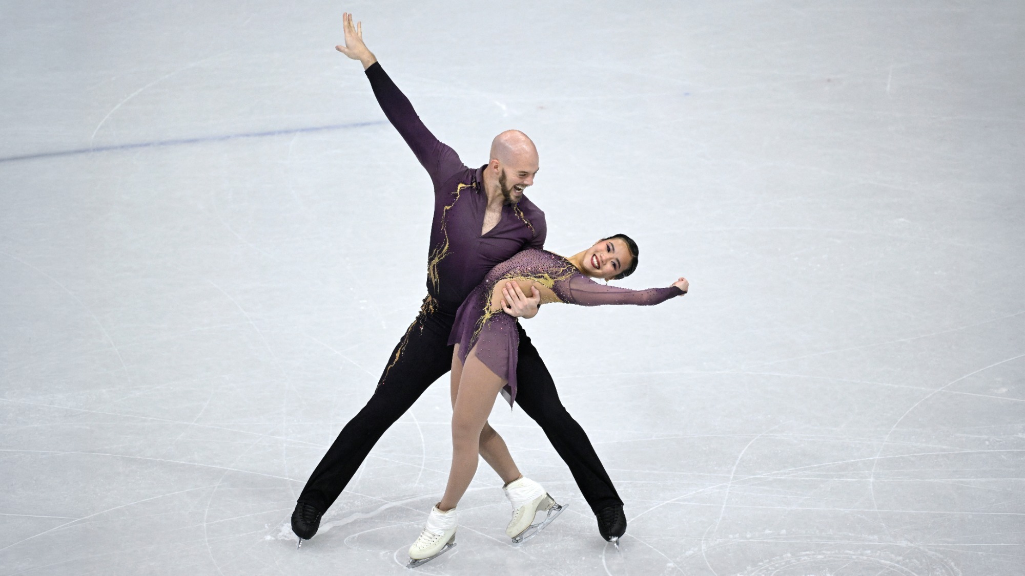Ellie Kam and Danny O'Shea hit their ending pose with triumphant smiles on their faces. Danny dips Ellie with the other hand extended upward. Ellie extends her left arm outward. Danny is a bald man wearing a purple long sleeve shirt with gold accents and black pants. Ellie is a woman wearing a matching long sleeve purple dress with gold accents and white skates. 