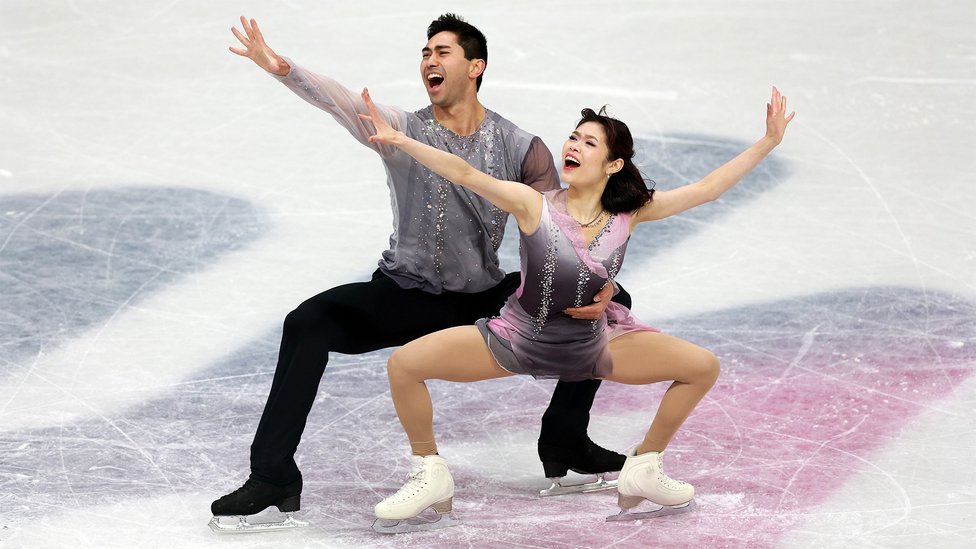  Emily Chan and Spencer Akira Howe skate together with Spencer skating behind Emily with his arm around her waist. His other arm and both of Emily's arms are extended. They both have elated looks on their faces. Emily is an Asian woman with short black hair wearing a purple skating costume. Spencer is an Asian man with short black hair wearing a matching purple long sleeve shirt and black pants.