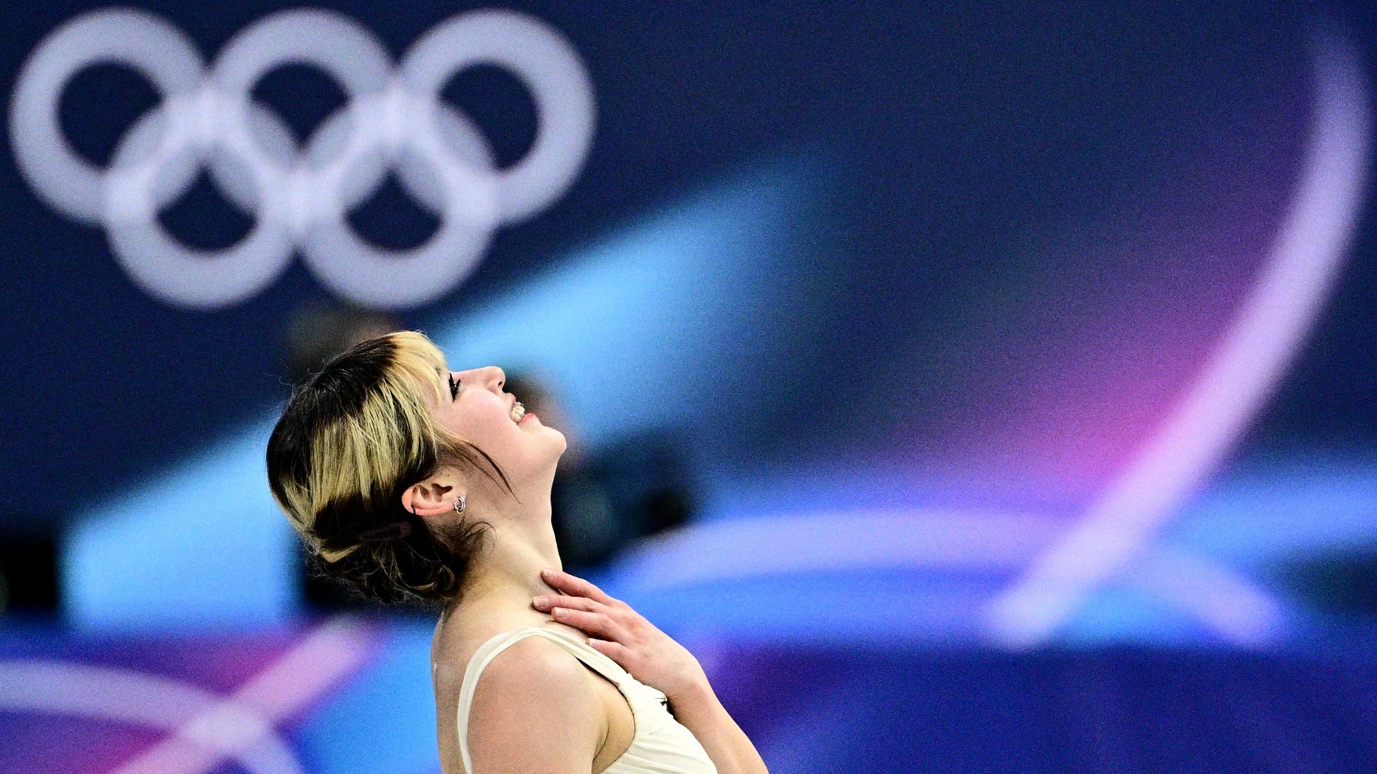 Alysa Liu smiles with one hand over her heart, looking upwards. She is a young woman with brown hair with blonde stripes wearing a white sleeveless skating costume. 