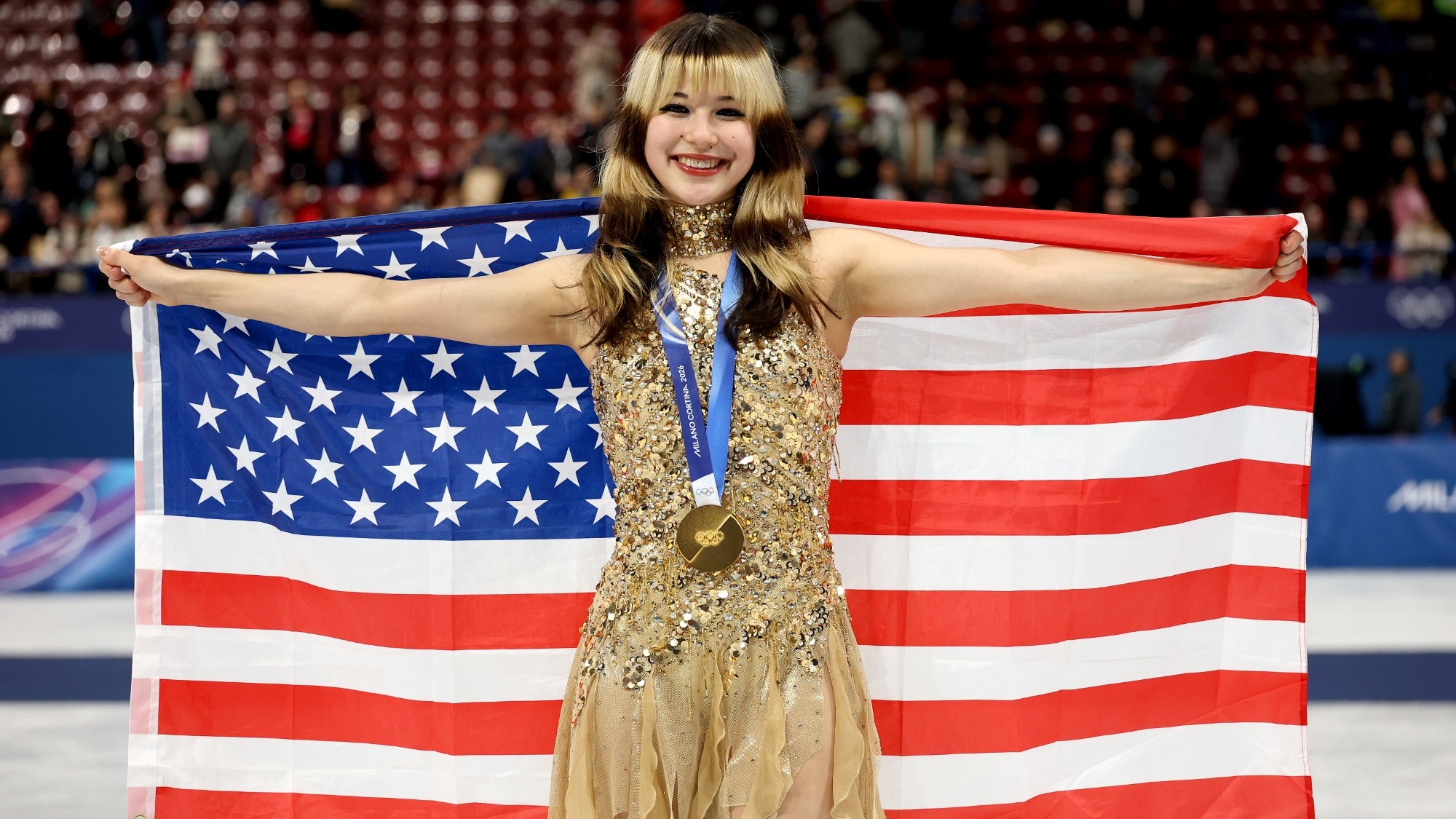 Alysa Liu stands smiling while holding an American flag behind her with an Olympic gold medal around her neck. Alysa is a young woman with brown hair with blonde horizontal stripes wearing a one-shoulder sparkly gold skating dress with a gold collar. 