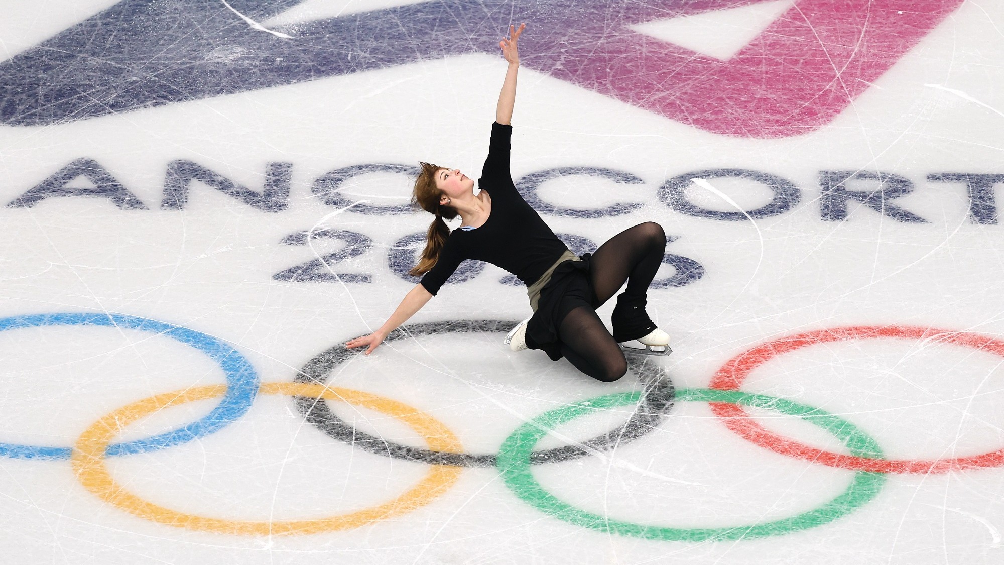 Alysa Liu performs a knee slide, looking up to the sky with one arm outstretched. Alysa is a young woman with brown hair tied back in a ponytail wearing a black long sleeve top and a black skirt over black pants. She skates over the Olympic rings and 2026 Olympic Winter Games logo on the ice