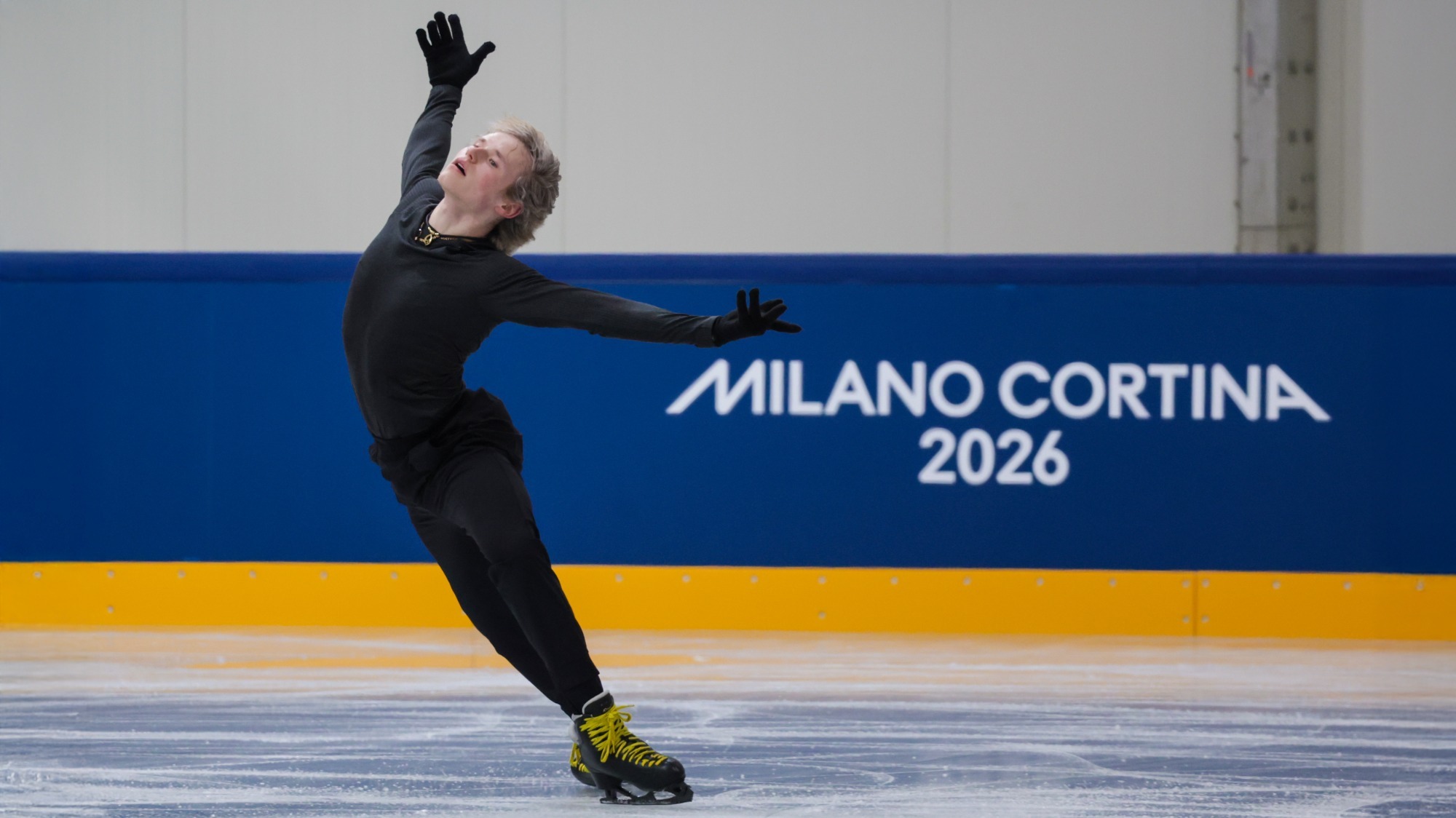 Ilia Malinin skates with his arms outstretched, looking up. Ilia is a young man with short blonde hair wearing a long sleeve black shirt, black pants and black skates with yellow laces. He skates in front of blue boards with the Olympic rings on them. 