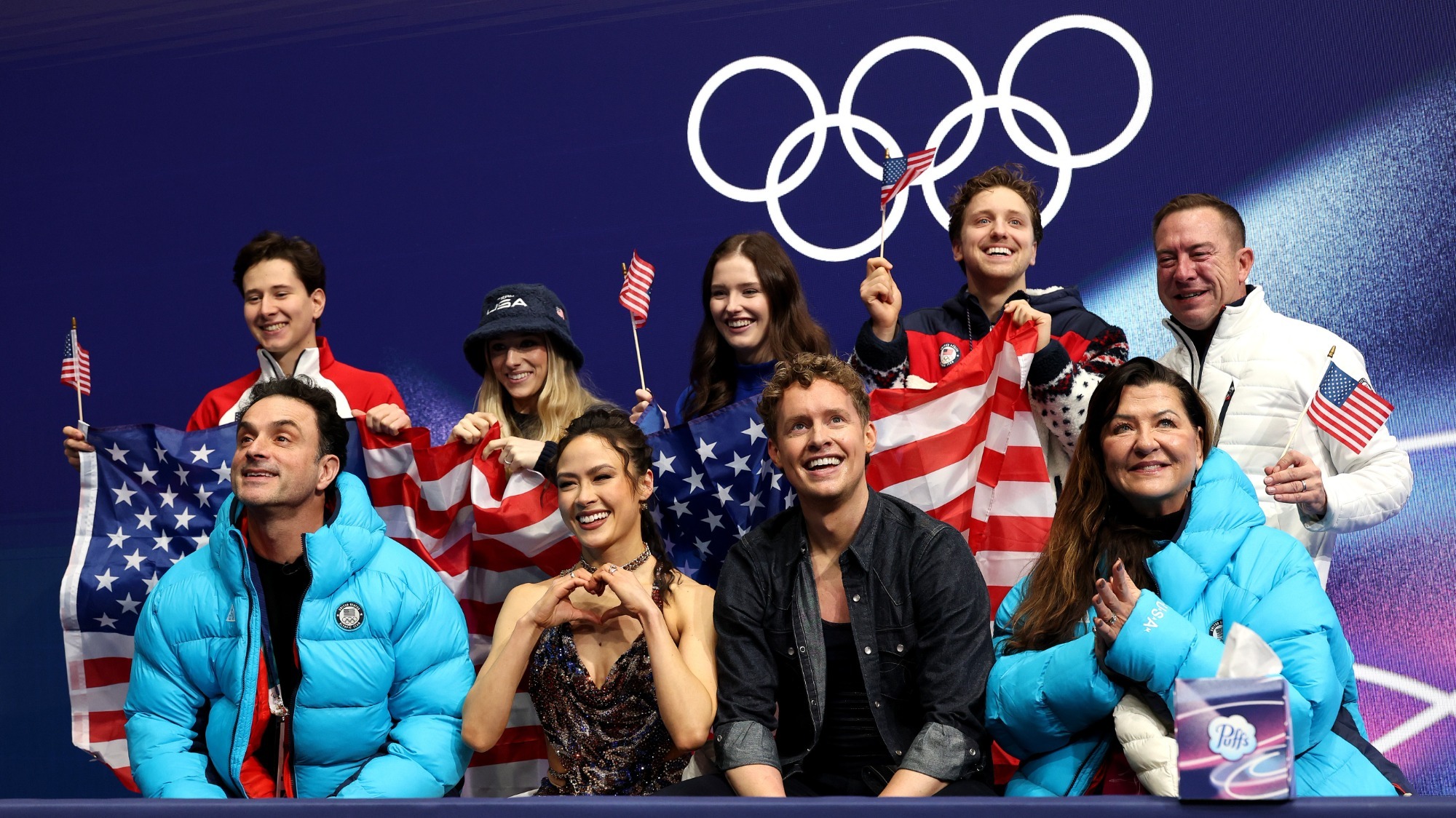 Madison Chock and Evan Bates sit in the team box during the Olympic team event waiting to receive their score. Both are smiling and Madison is making a heart with her hands. Their coaches stand on either side of them and other members of Team USA stand behind them cheering and holding American flags. The Olympic rings are on the wall in the background