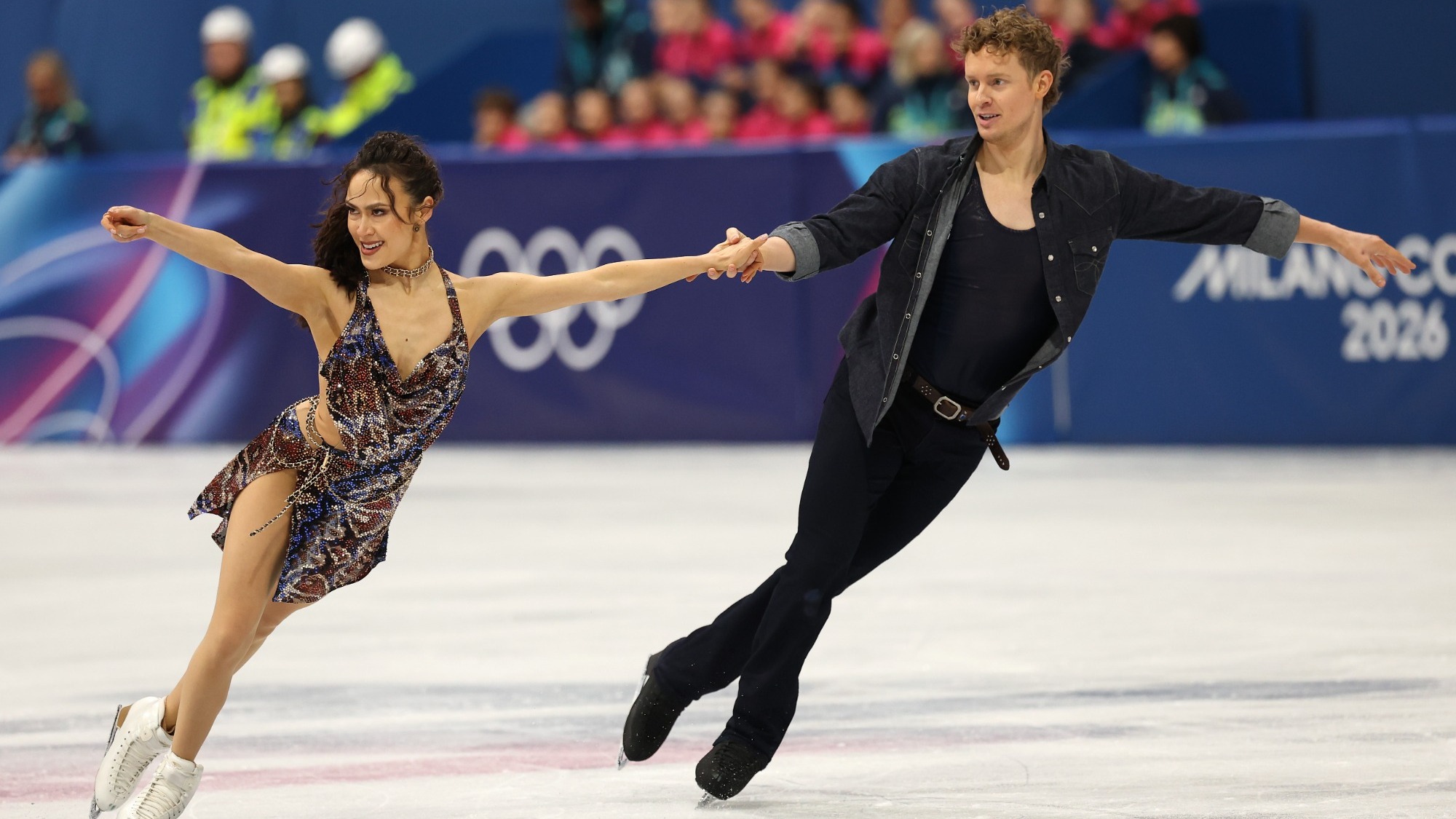 Madison Chock and Evan Bates skate hand in hand looking to the left. Madison (left) is a woman with long dark hard wearing an animal print dress. Evan (right) is a tall man with curly blonde hair wearing a black denim button down jacket over a black shirt with black pants. 