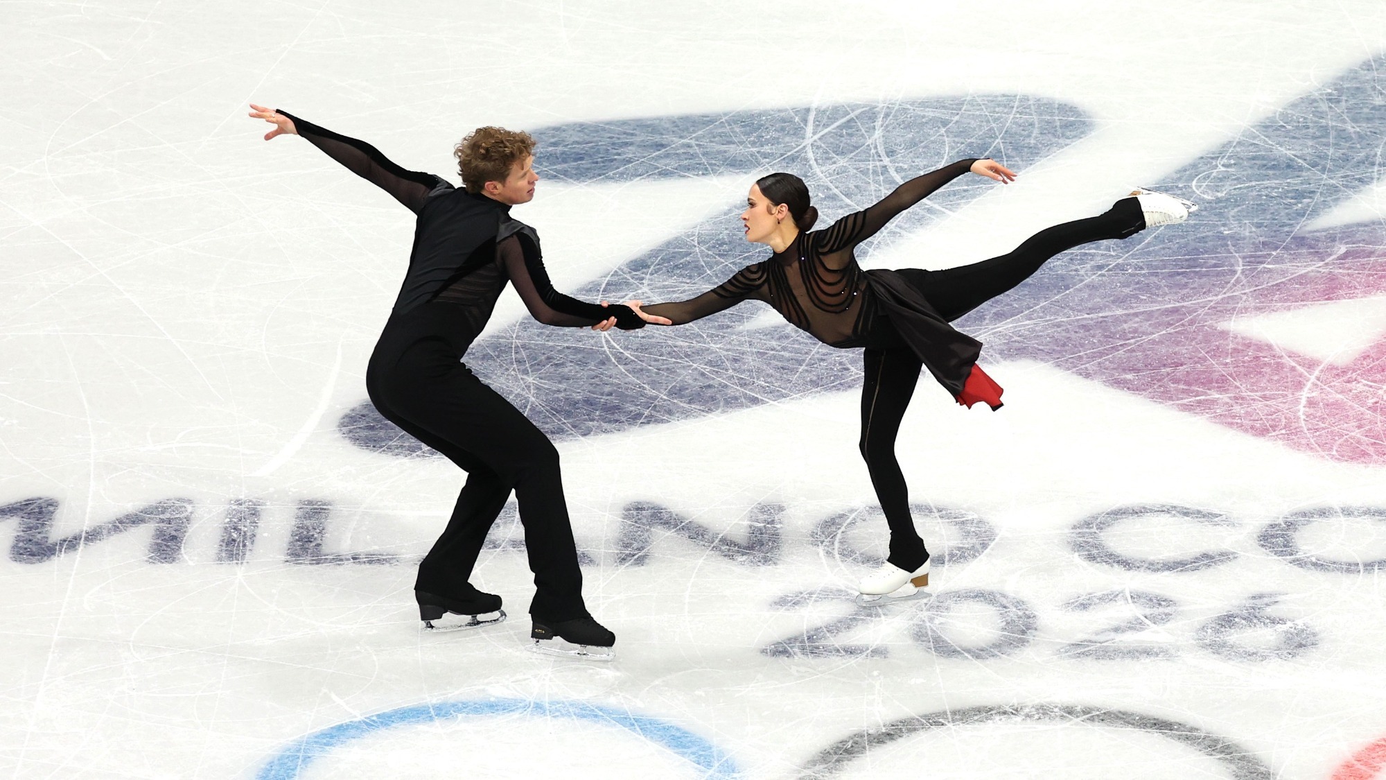 Madison Chock and Evan Bates skate facing each other, holding each others hand with one leg extended outward. Evan (left) is a man with short curly blonde hair wearing a black long sleeve top and black pants. Madison is a woman with dark hair tied back in a low bun wearing a black long sleeve skating costume with a long black skirt with red on the inside. 