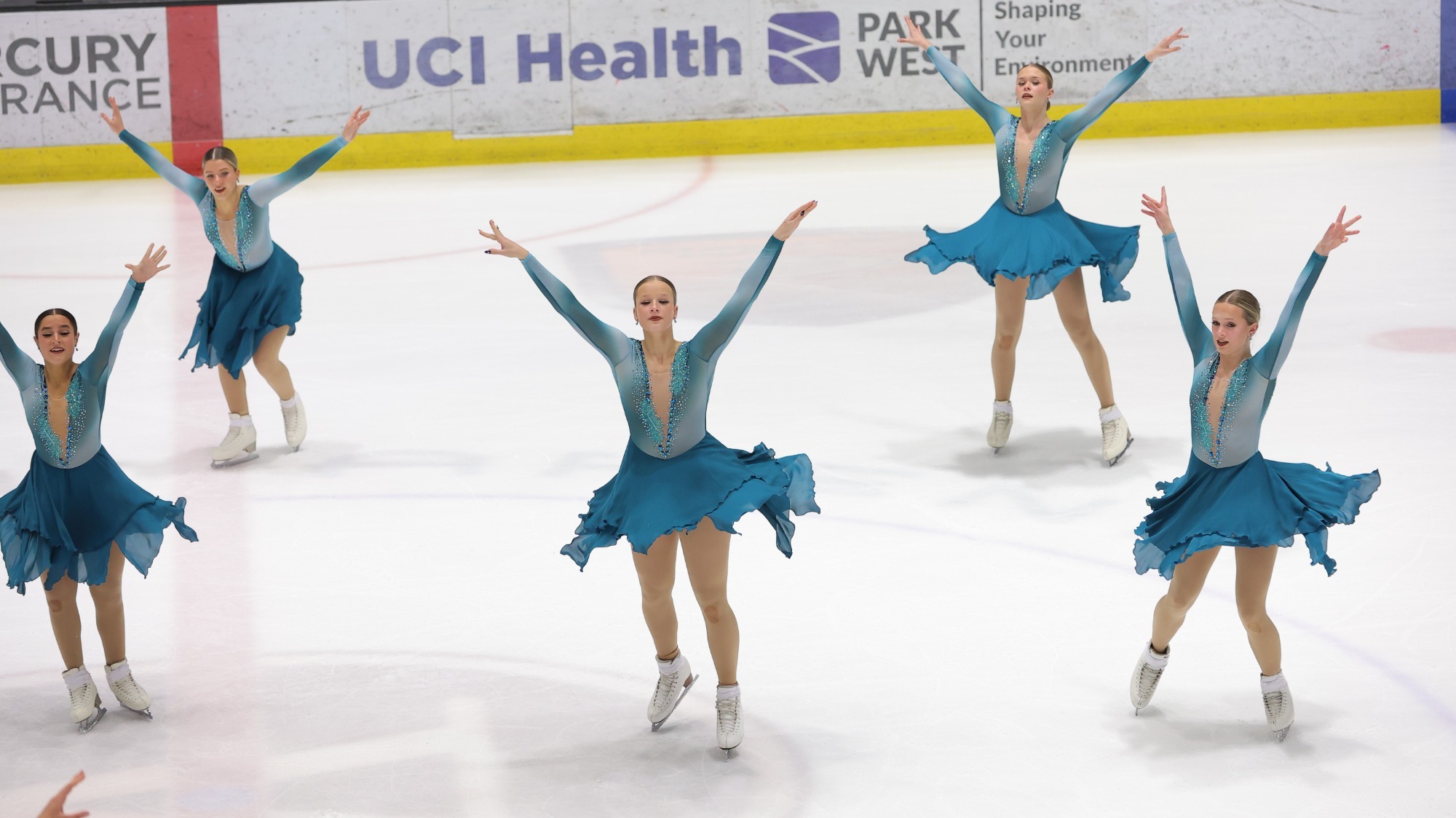 The Northernettes skate with their arms stretched upward in unison. All team members are wearing matching teal long sleeve skating costumes with a coordinating teal skirt. 