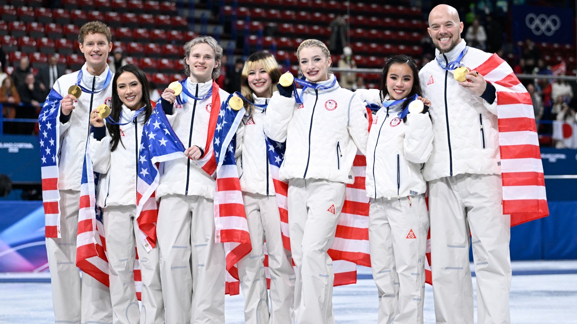 Team USA stands holding their gold medals and smiling wearing matching white jackets and white pants. The athletes stand with American flags draped around their shoulders. 