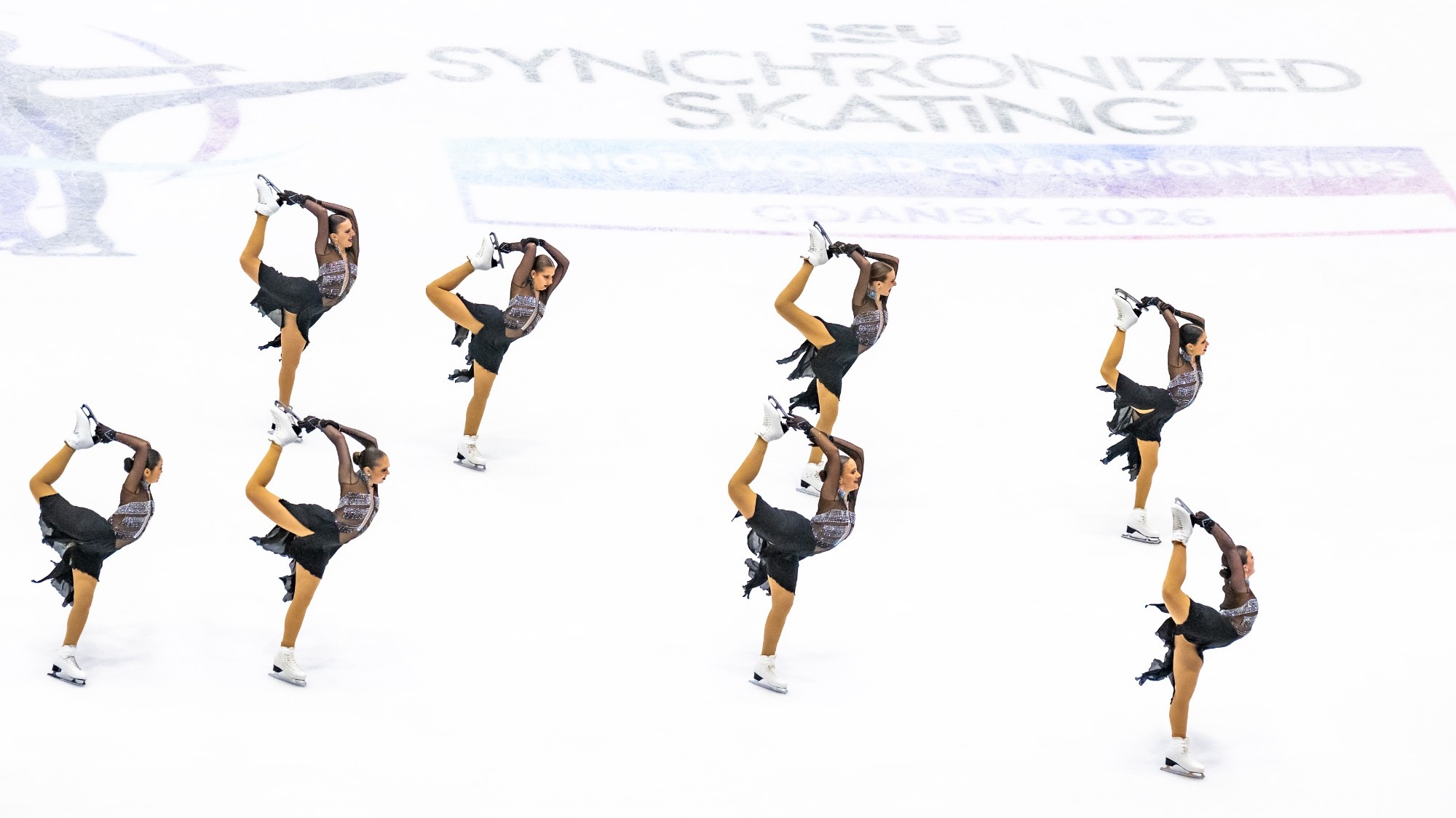 Skyliners junior, wearing black dresses with silver detail on the top, skate across the center ice logo at Junior Worlds. The eight skaters are all holding their back leg above their head as they skate.