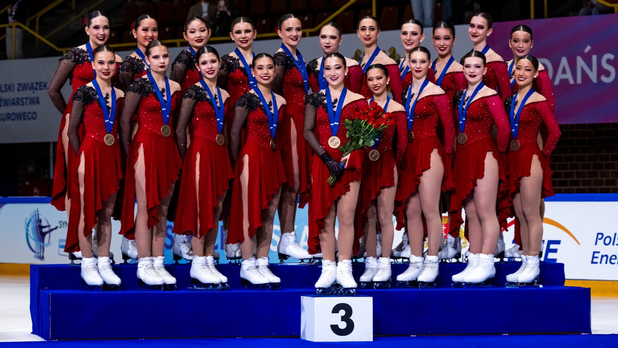 Skyliners junior stand on the podium with bronze. They all wear red dresses with black detail on the left shoulder, bronze medals around their necks and smile.