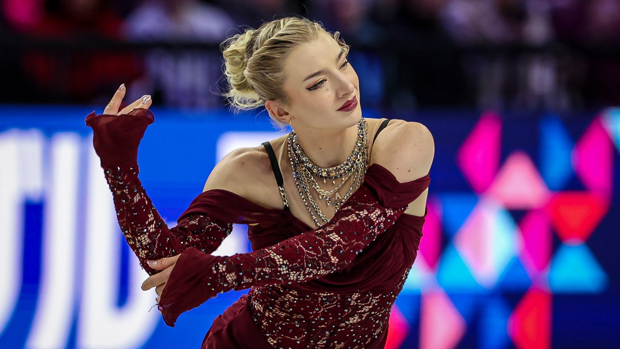 Photo taken from the waist up of Amber Glenn looking to the right with her arms extended. Amber is a woman with blonde hair tied up in a bun wearing a maroon skating costume with lace sleeves and midsection and chunky gold necklaces around her neck. 