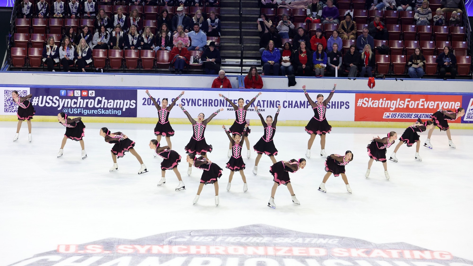 Teams Elite juvenile stands in a triangle formation wearing matching black skating costumes with pink ribbon detailing. 
