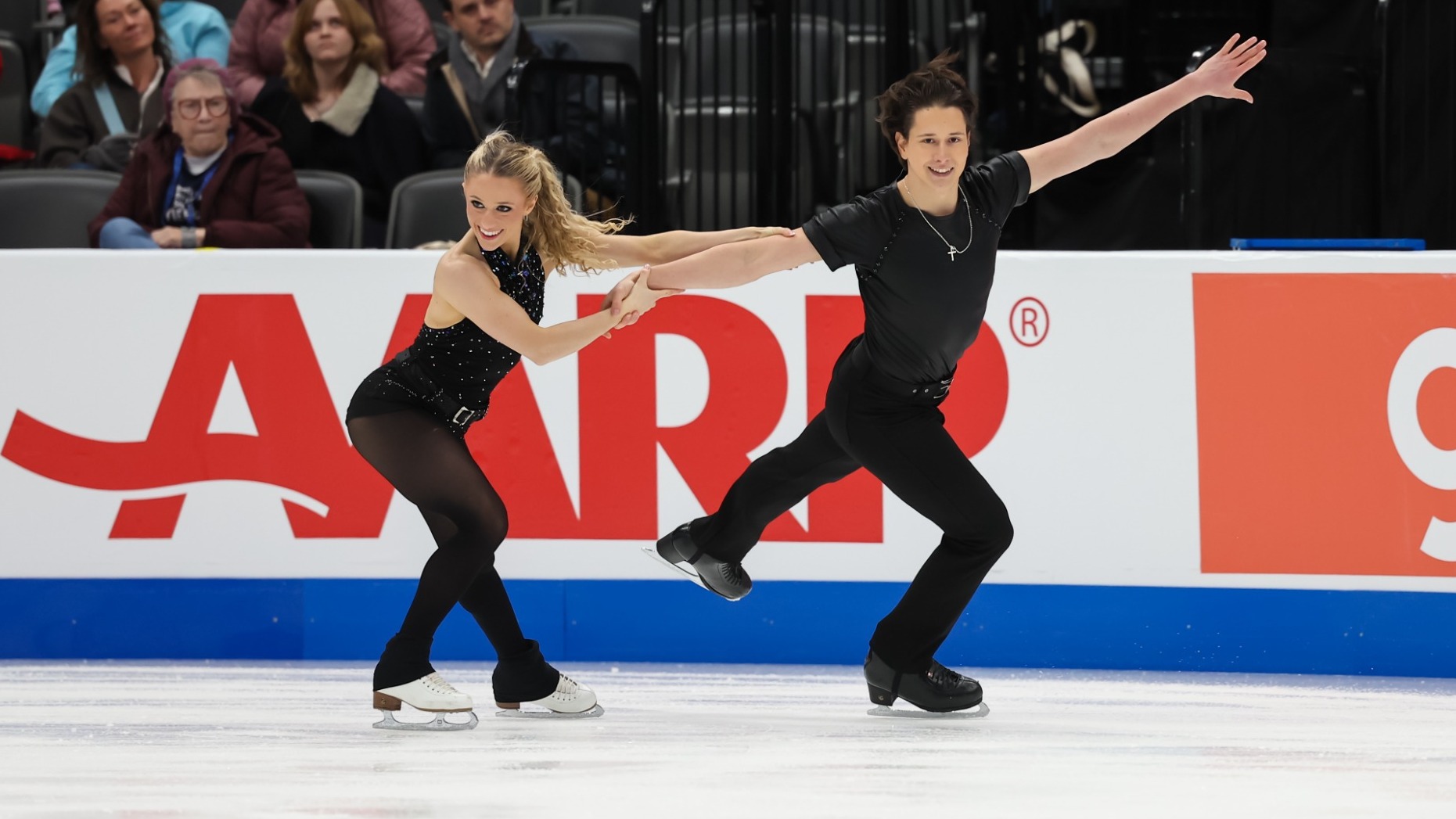 Emilea Zingas and Vadym Kolesnik skate side by side with Emilea holding on to Vadym's outstretched hand. Emilea is a young woman with long blonde hair wearing a sparkly sleeveless top and black pants. Vadym is a young man with short black hair wearing a black tshirt and black pants. 