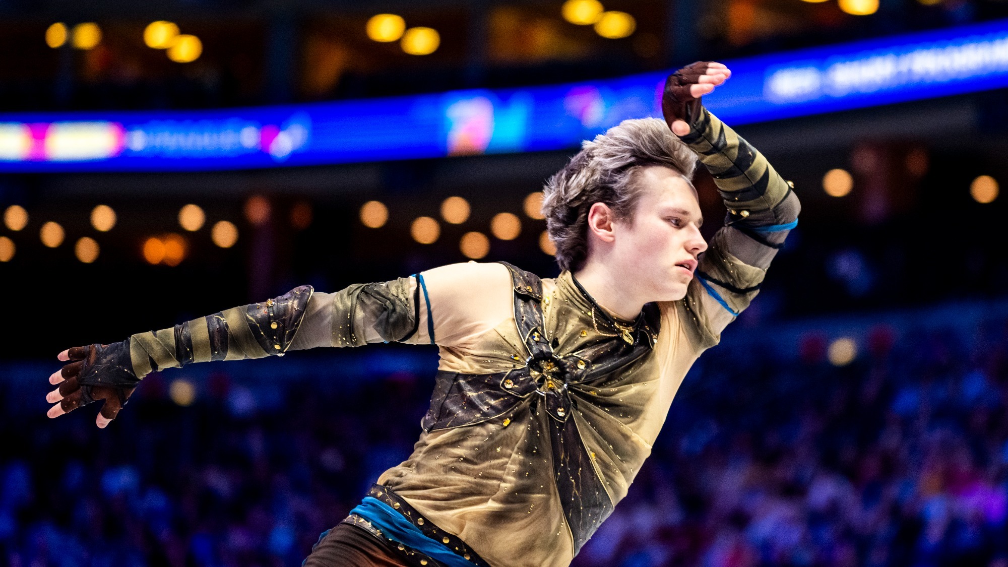 Photo taken from the waist up of Ilia Malinin skating with hand up by his face and the other extended outward. Ilia is a young man with short blonde hair Ilia is a young man with short blonde hair wearing a brown long sleeve top with jewel details, matching brown gloves a blue belt and matching brown pants.
