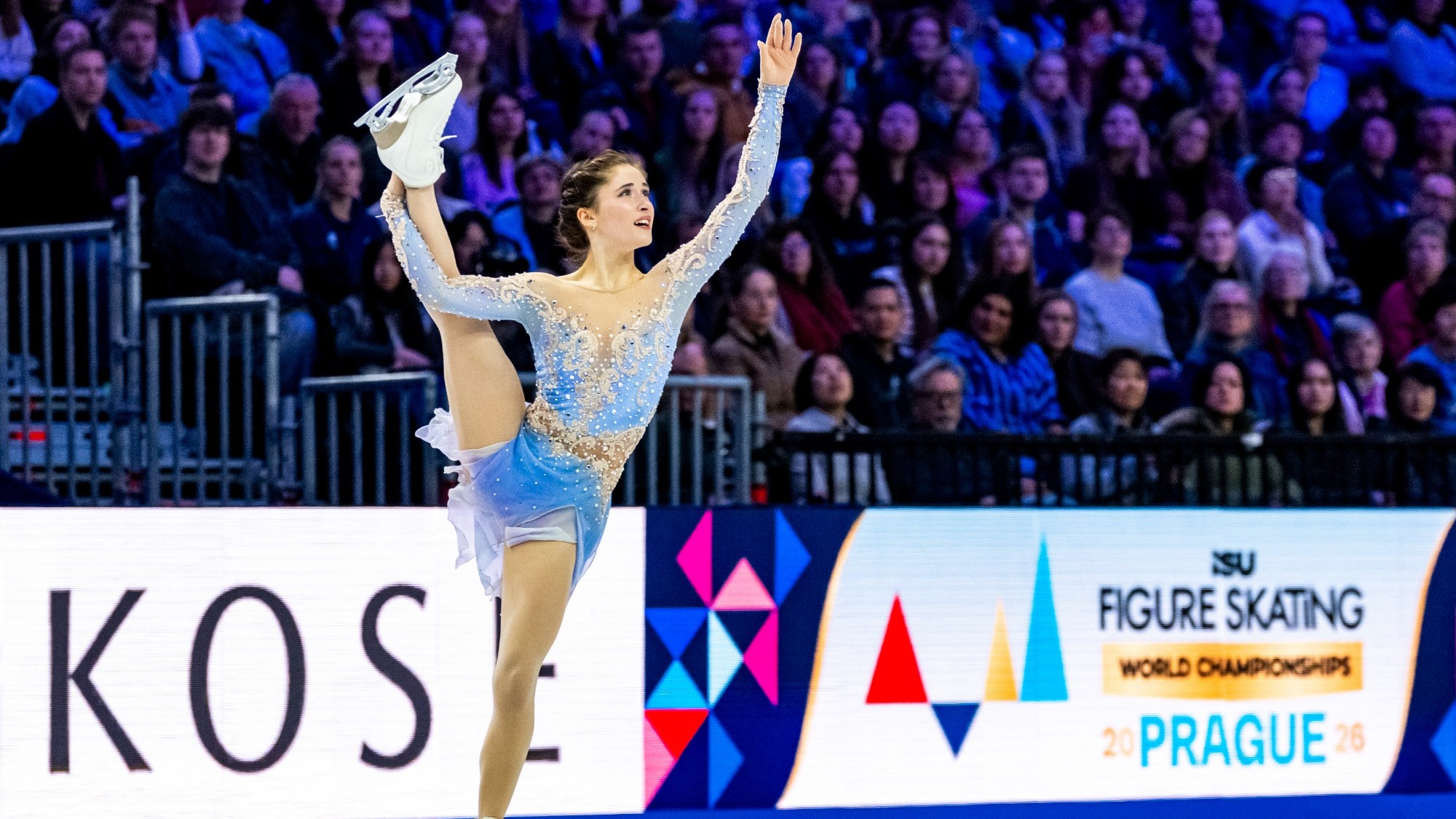 Isabeau Levito skates during the women's free skate at the 2026 World Championships. She skates holding one leg above her head. Isabeau is a young woman with brown hair tied up in a bun wearing a long sleeve light blue skating costume with white crustal detailing. A full crowd of fans can be seen behind her. 