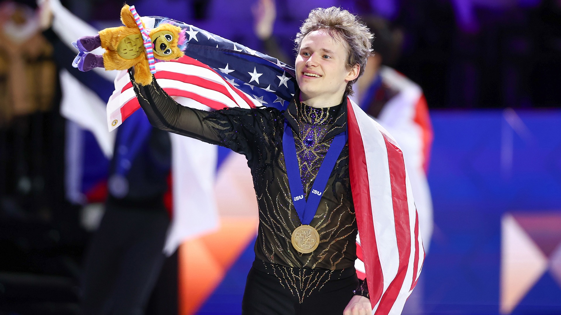 Ilia Malinin skates a victory lap waving to the crowd with a smile on his face. He holds an American flag behind him and a stuffed hedgehog toy in his right hand. Ilia is a young man with blonde hair wearing a long sleeve skating costume with gold and blue jewel detailing down the front. He is also wearing matching black