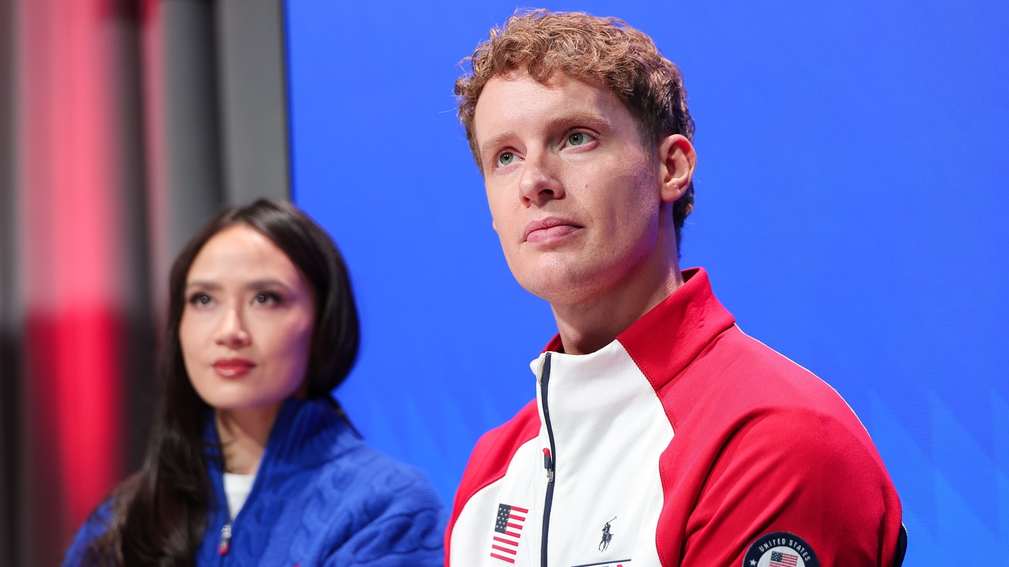 Photo taken from the waist up of Evan Bates sitting in a panel discussion looking to the left. Evan is a man with short curly blonde hair wearing a jacket with a white torso and red sleeves