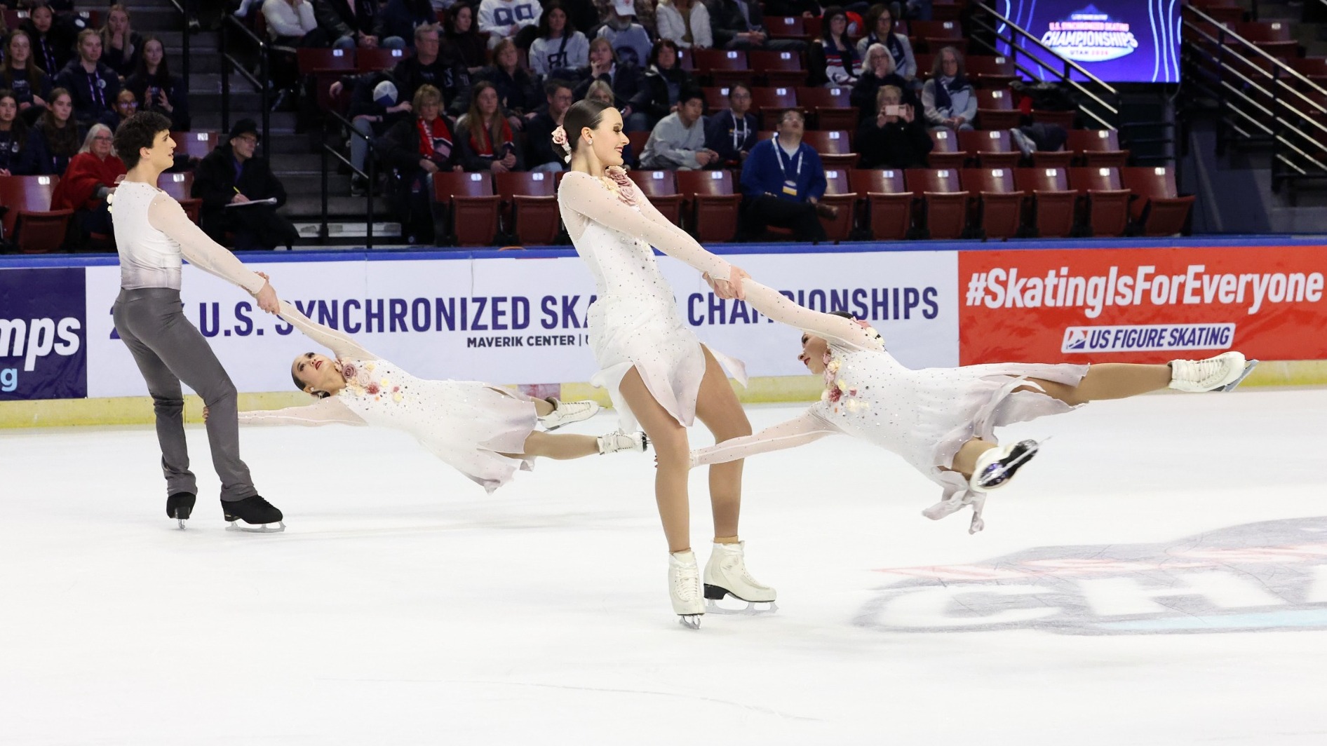 Teams Elite junior performs an element with one skater being swung around by their teammate. Female skaters are wearing a white long sleeve dress with pink flower details. One male skater is wearing a matching top with grey pants